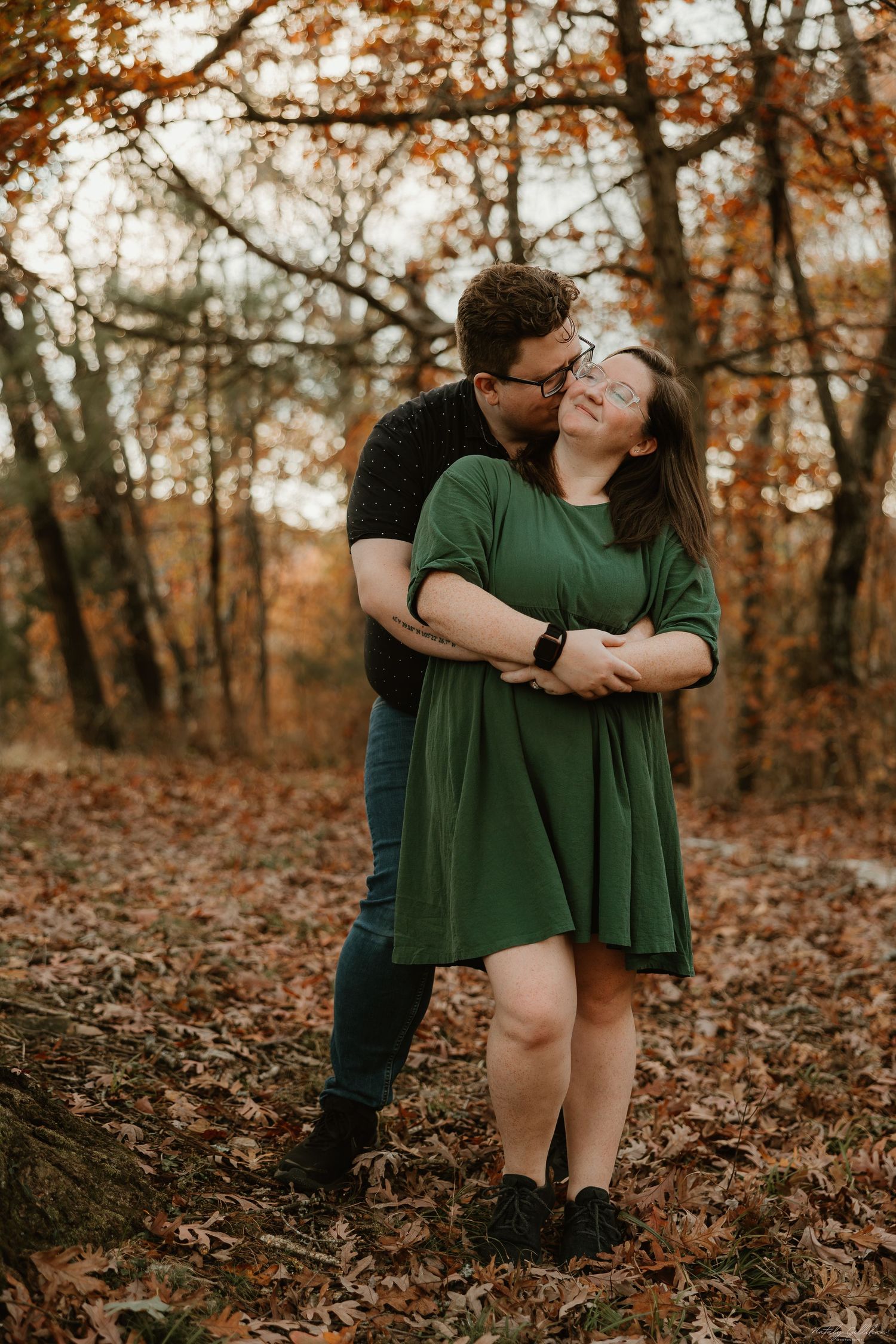 A couple shares an intimate moment in a forest during autumn with fallen leaves and warm autumn colors in the background.