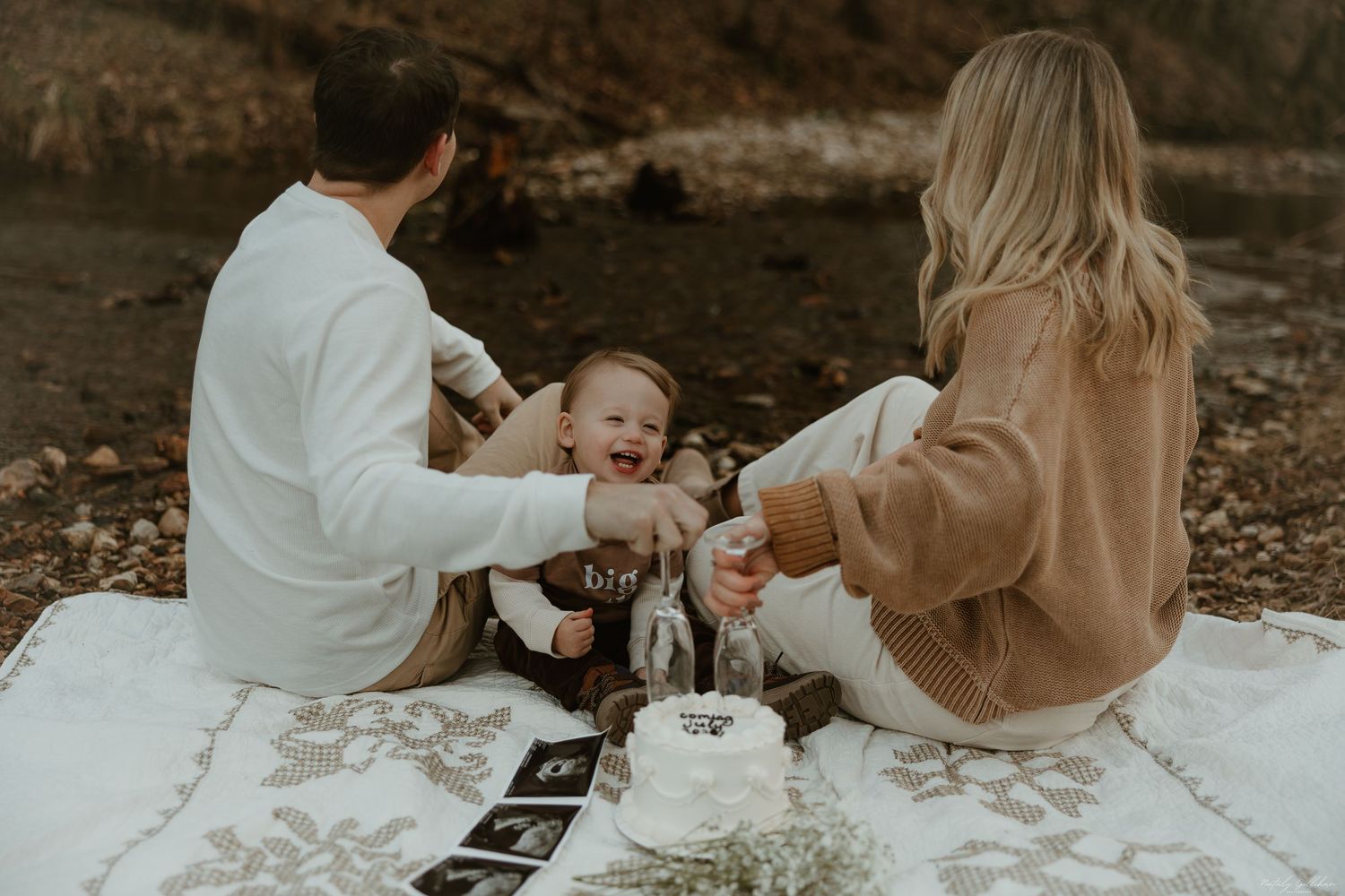 A family sits on a blanket having a picnic in a wooded area during autumn with a sonogram photo nearby.