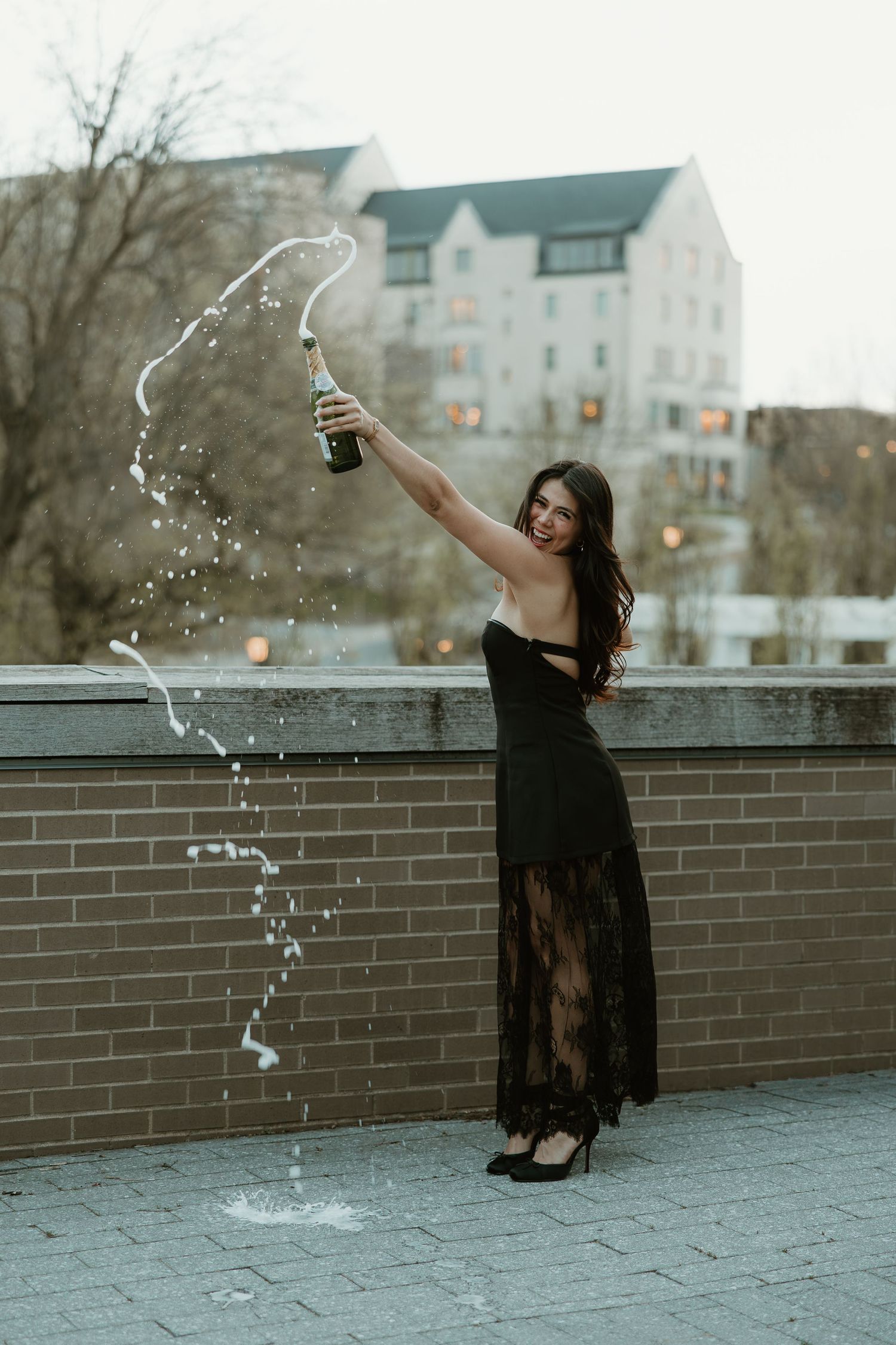 Woman in black lace gown popping champagne bottle on rooftop terrace, celebrating with bubbly splashing down.