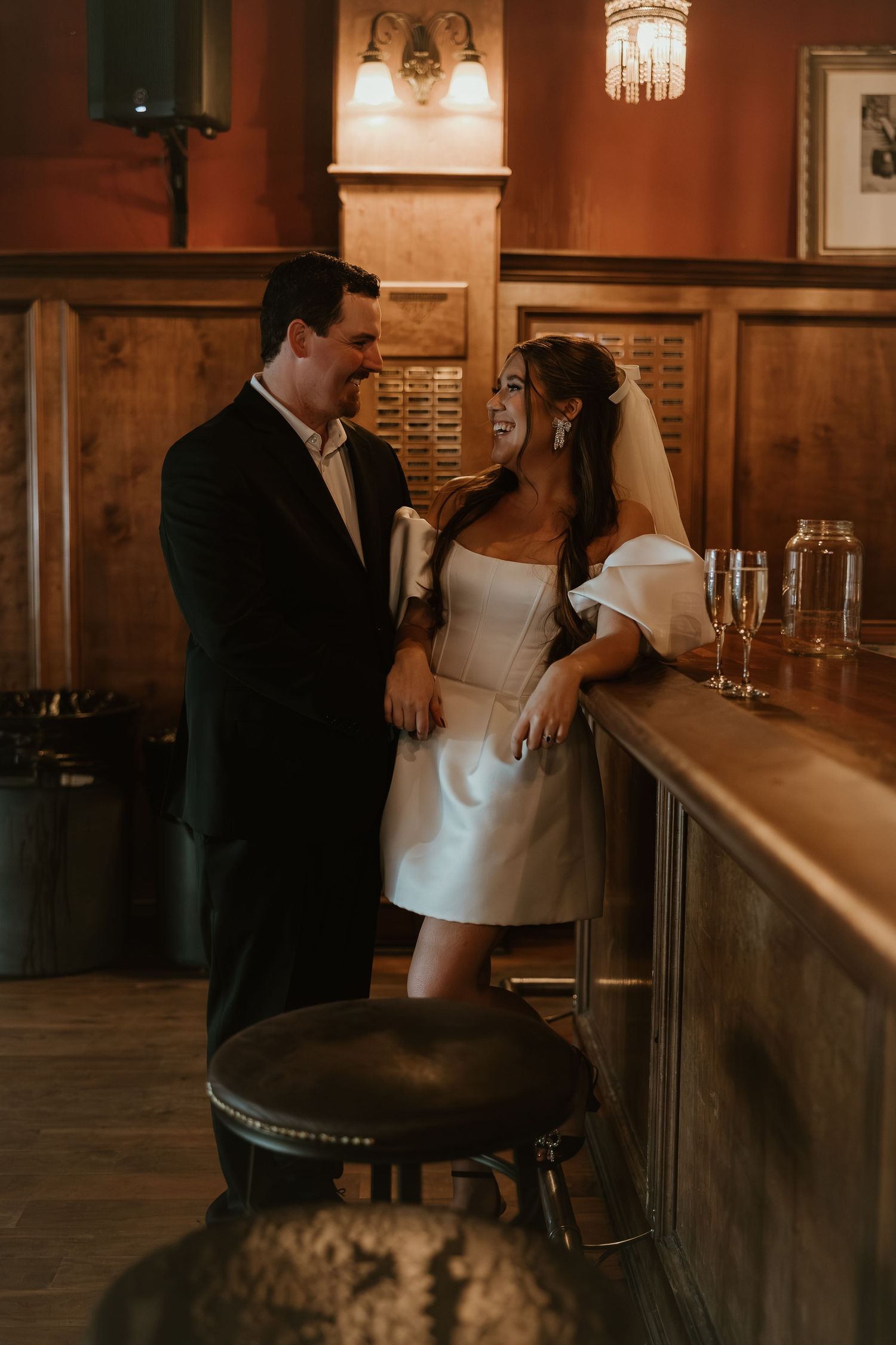 Bride in short white dress and groom in dark suit share a romantic moment at a warmly lit wooden bar.