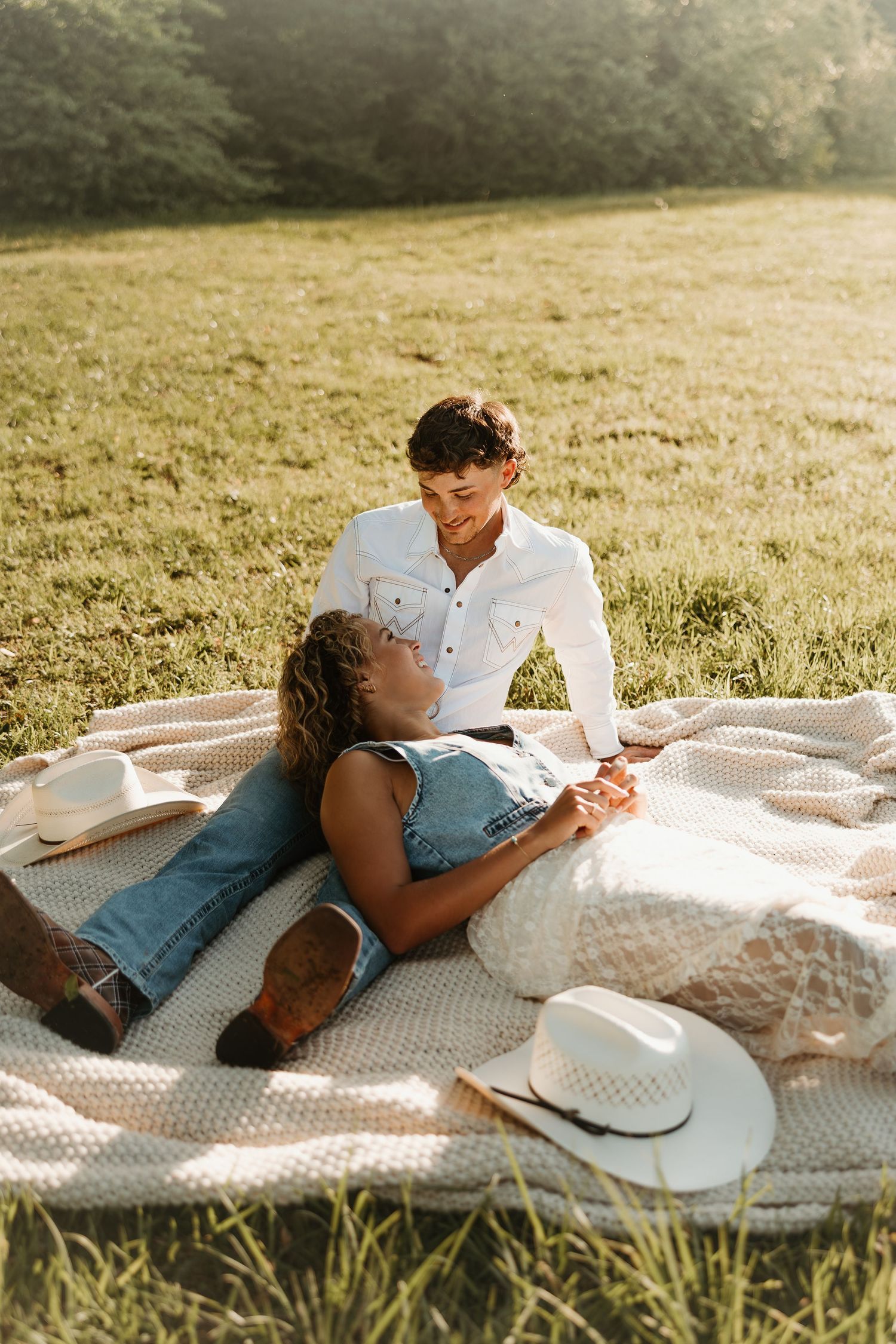 A couple shares an intimate moment while relaxing on a white blanket in a sunlit meadow at golden hour.