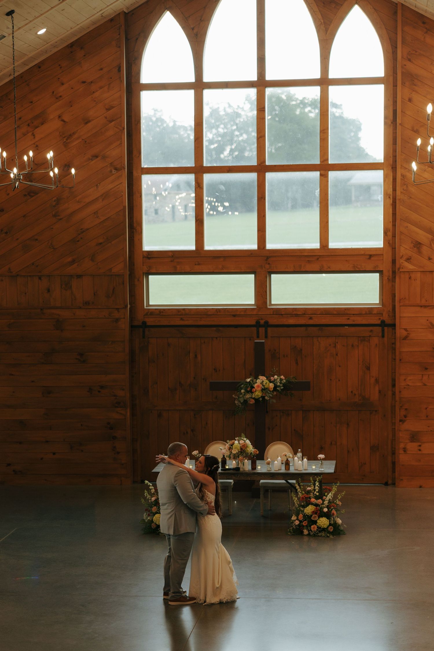 A couple shares a romantic dance in a rustic wooden barn venue with cathedral windows and warm lighting.