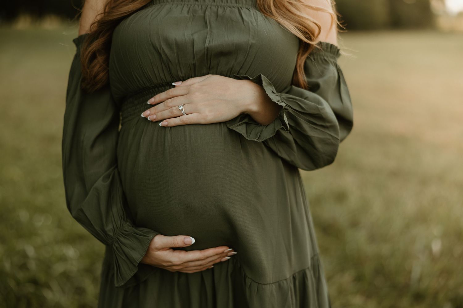 A maternity photoshoot shows a person in an olive green dress cradling their baby bump in a sunlit outdoor setting.