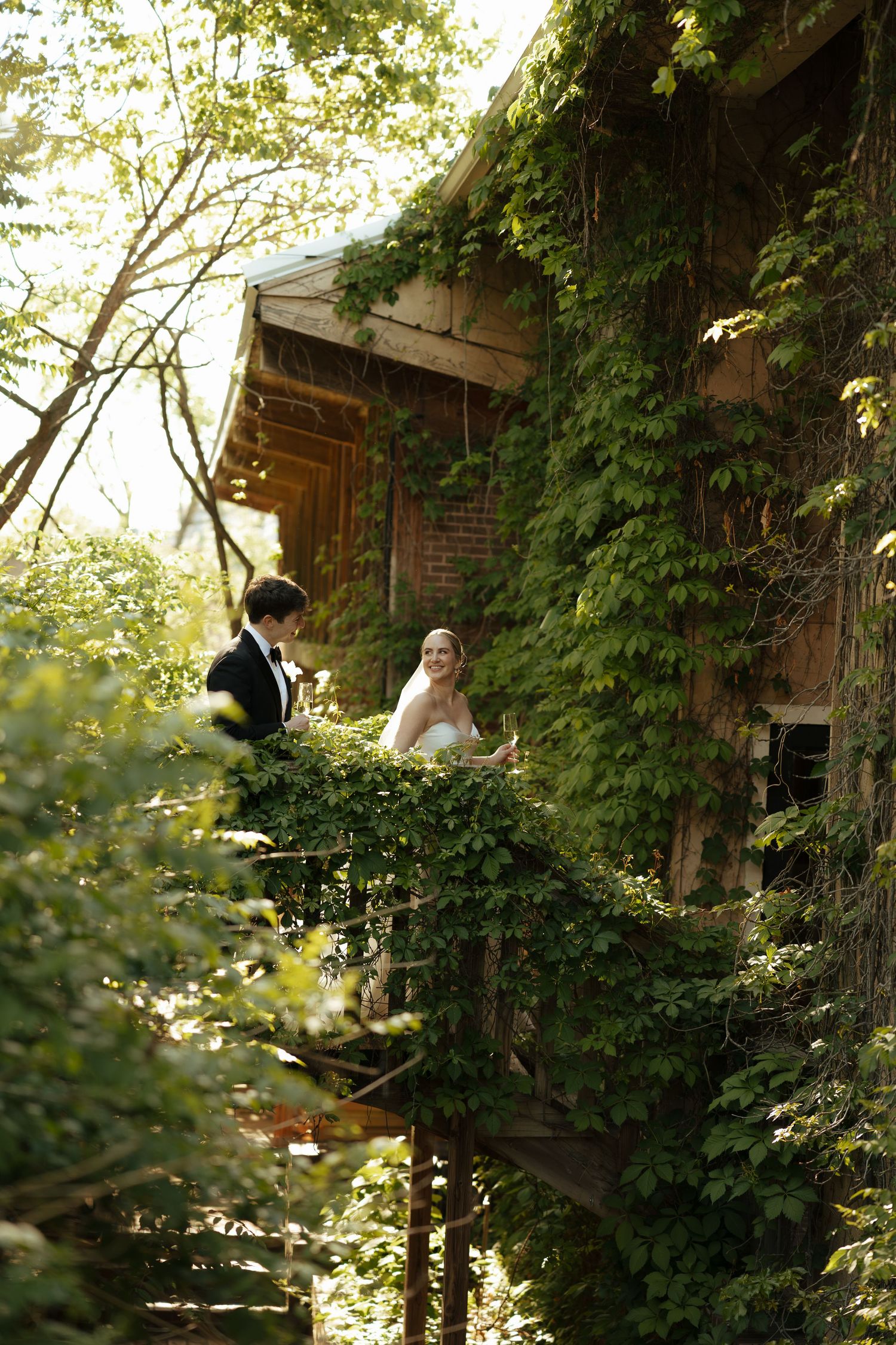 Bride and groom pose on an ivy-covered rustic building balcony surrounded by lush green trees.