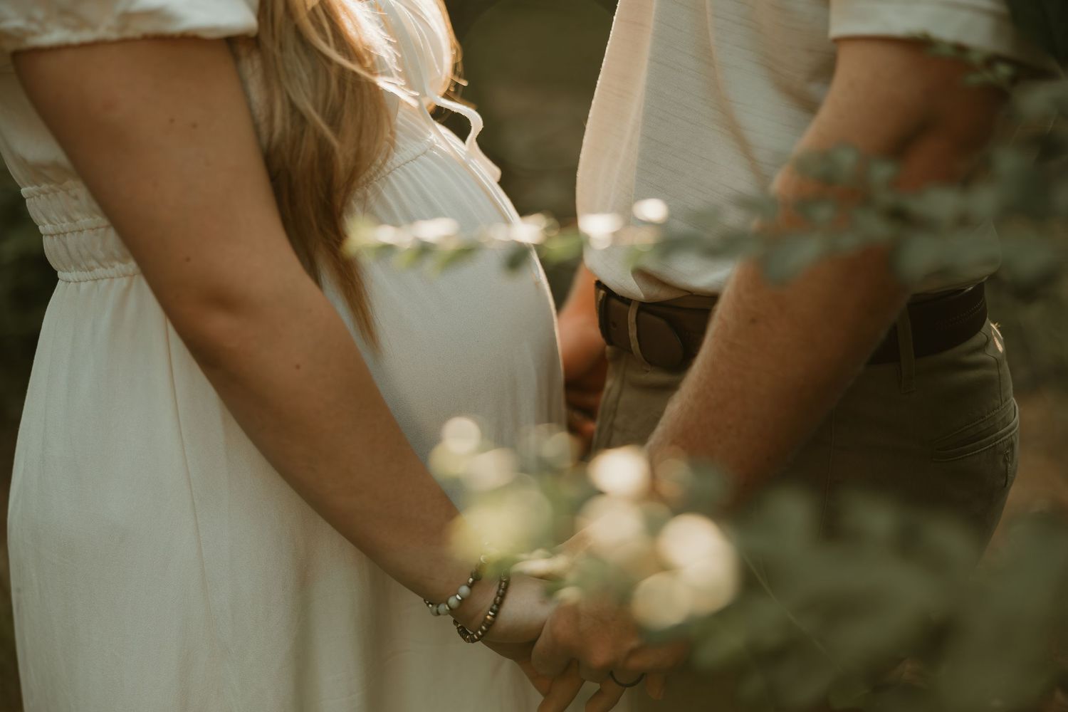Two people hold hands in an intimate moment wearing white and khaki clothing in a soft natural setting.