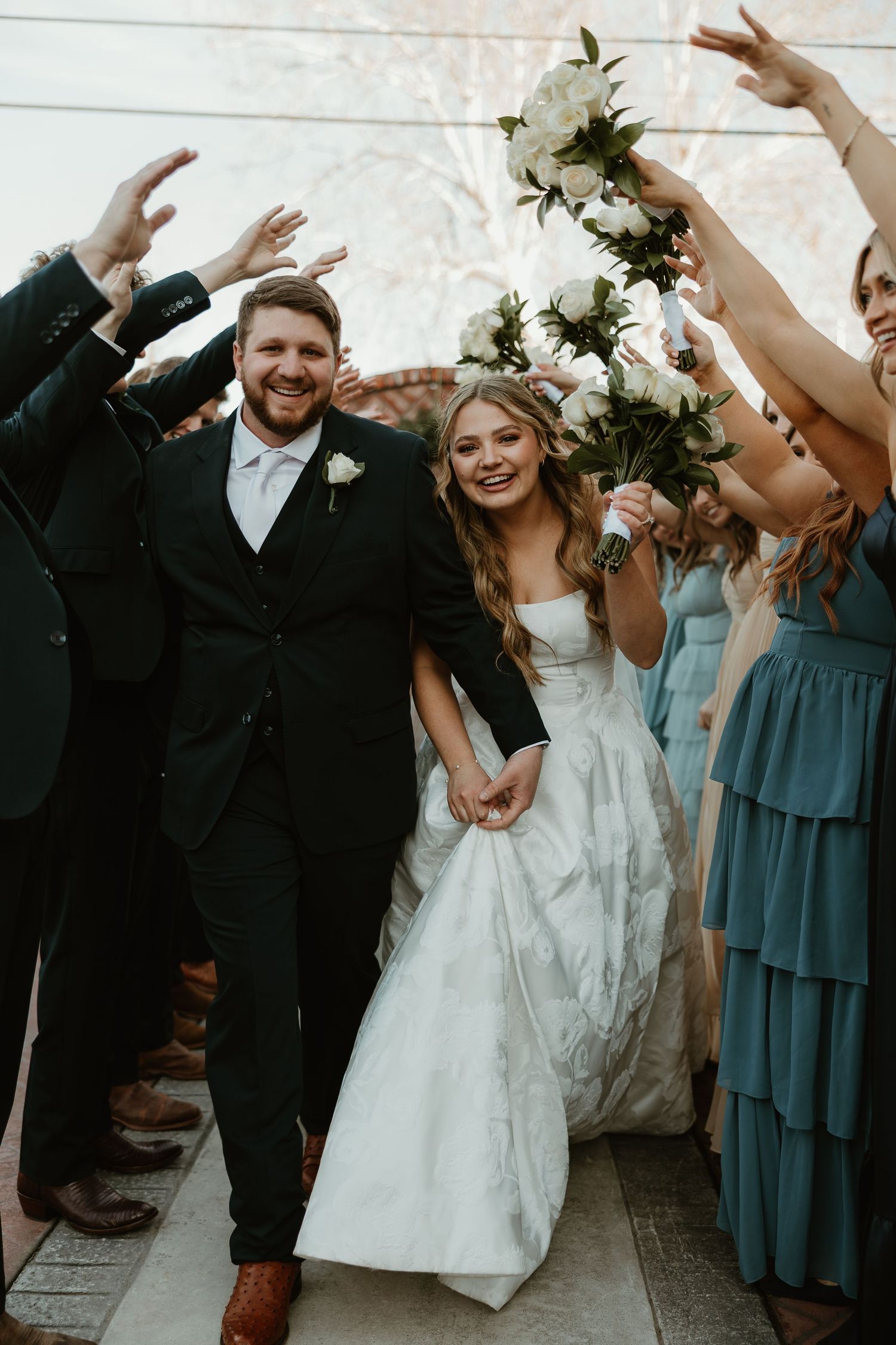 Wedding party celebrates with raised hands and white floral bouquets while wearing formal attire at outdoor venue.