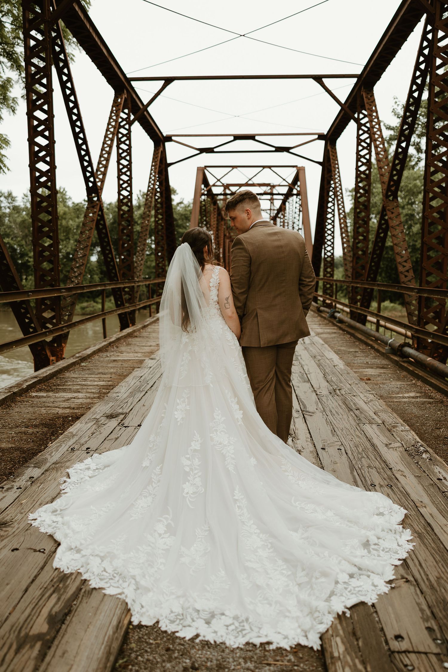 A bride in a lace train wedding dress and groom stand on a rustic wooden bridge with metal trusses.