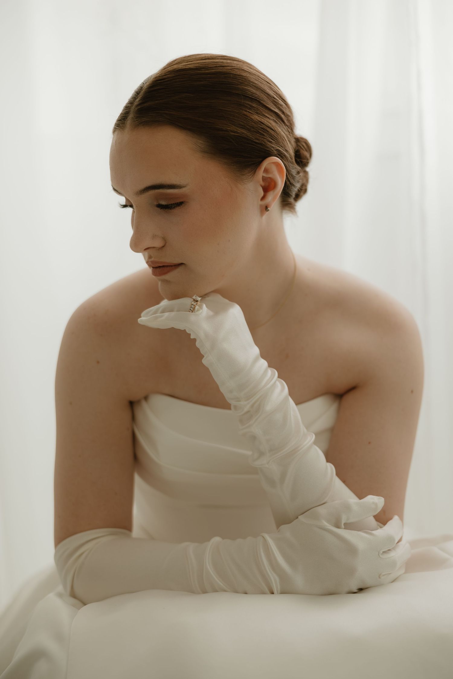 Elegant bride in white strapless gown and long satin gloves poses gracefully against soft white curtain backdrop.