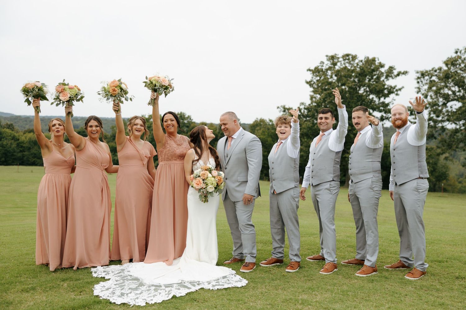 Wedding party in peach bridesmaid dresses and light gray suits pose together on grassy lawn during outdoor celebration.