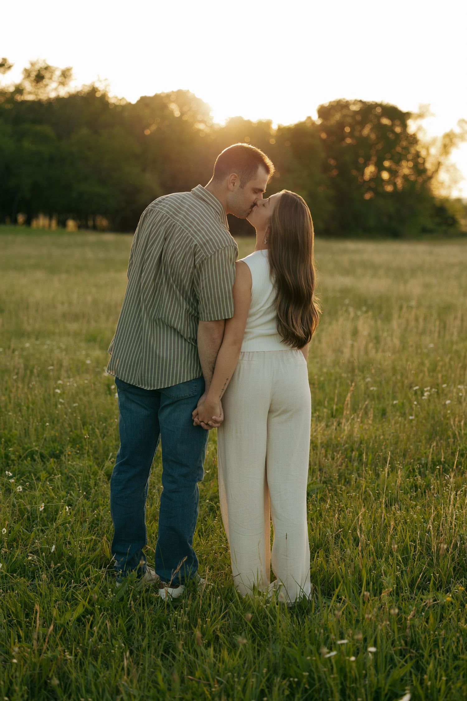 Couple kissing at golden hour in a green field, woman in white outfit holding hands with man in striped shirt and jeans.