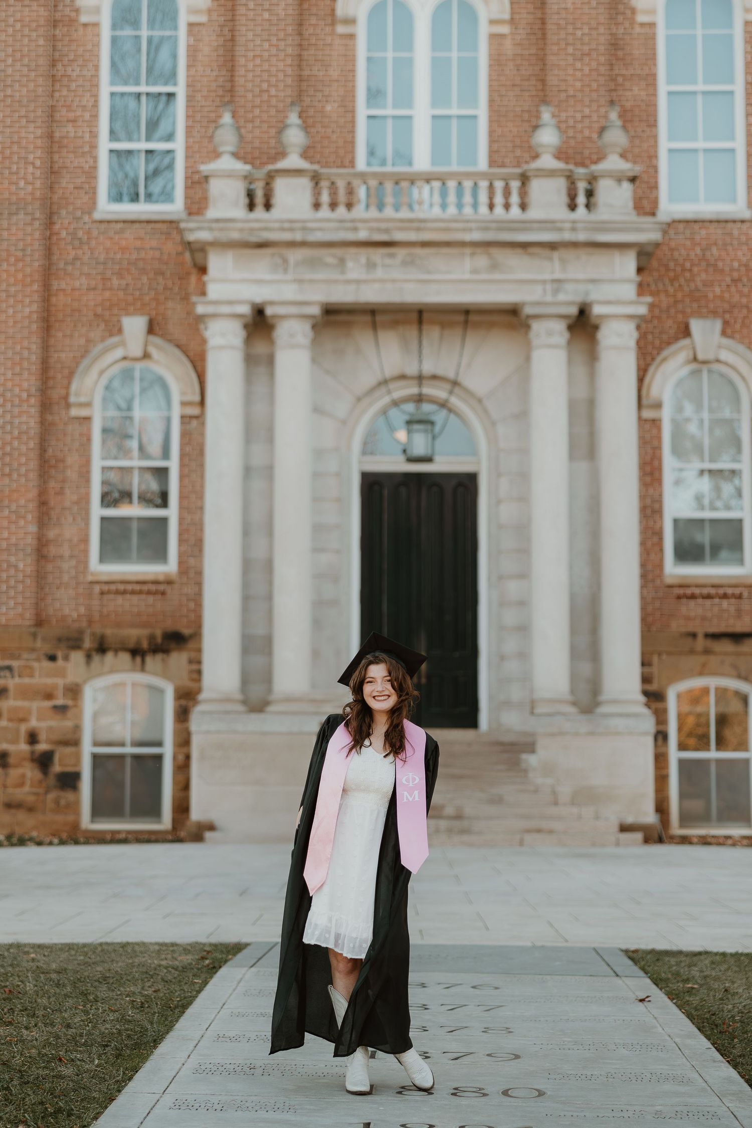 A person wearing a graduation cap and gown stands in front of a grand brick university building with arched windows.