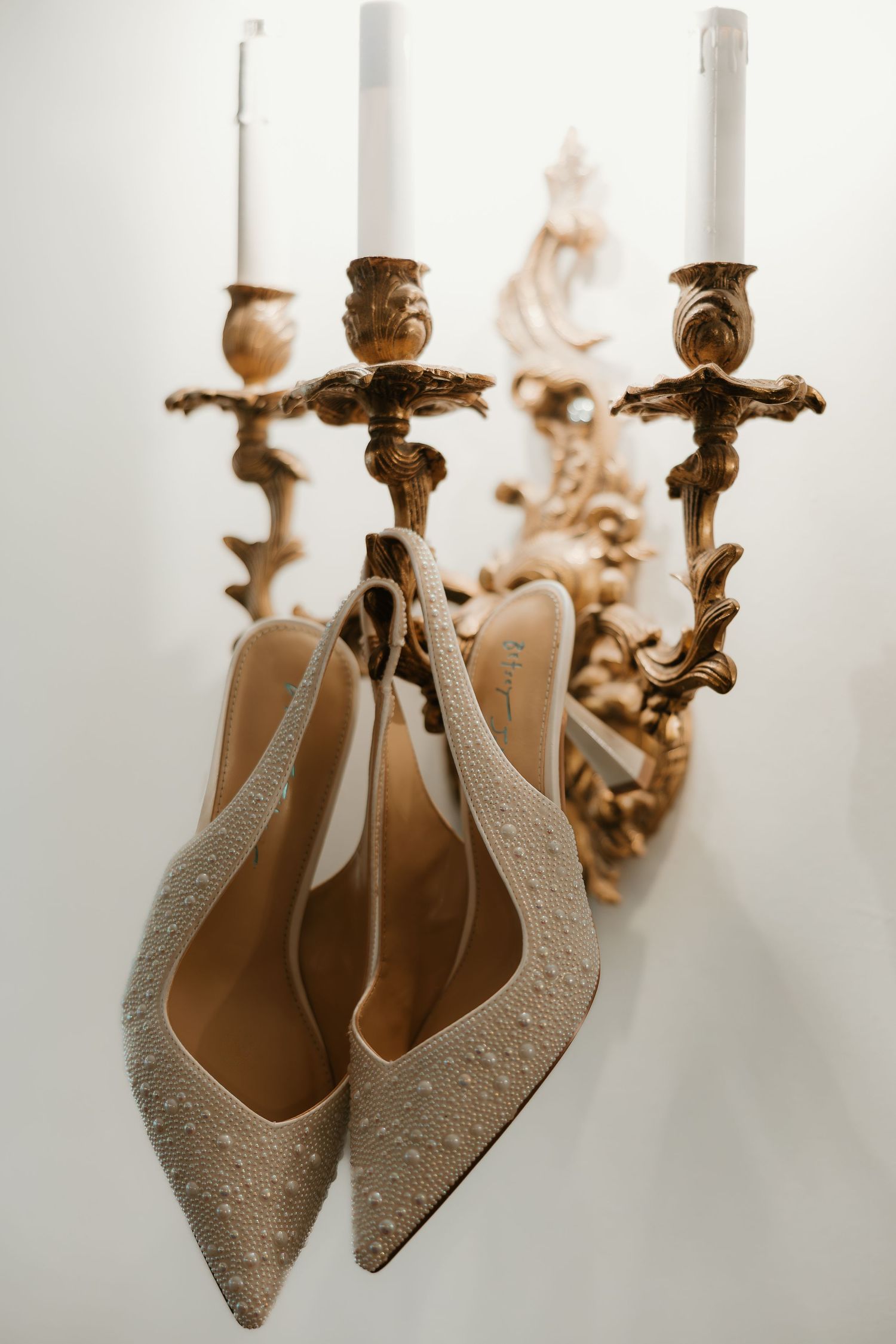 Elegant beige bridal shoes with embellishments hanging from an ornate gold candelabra against a white background.