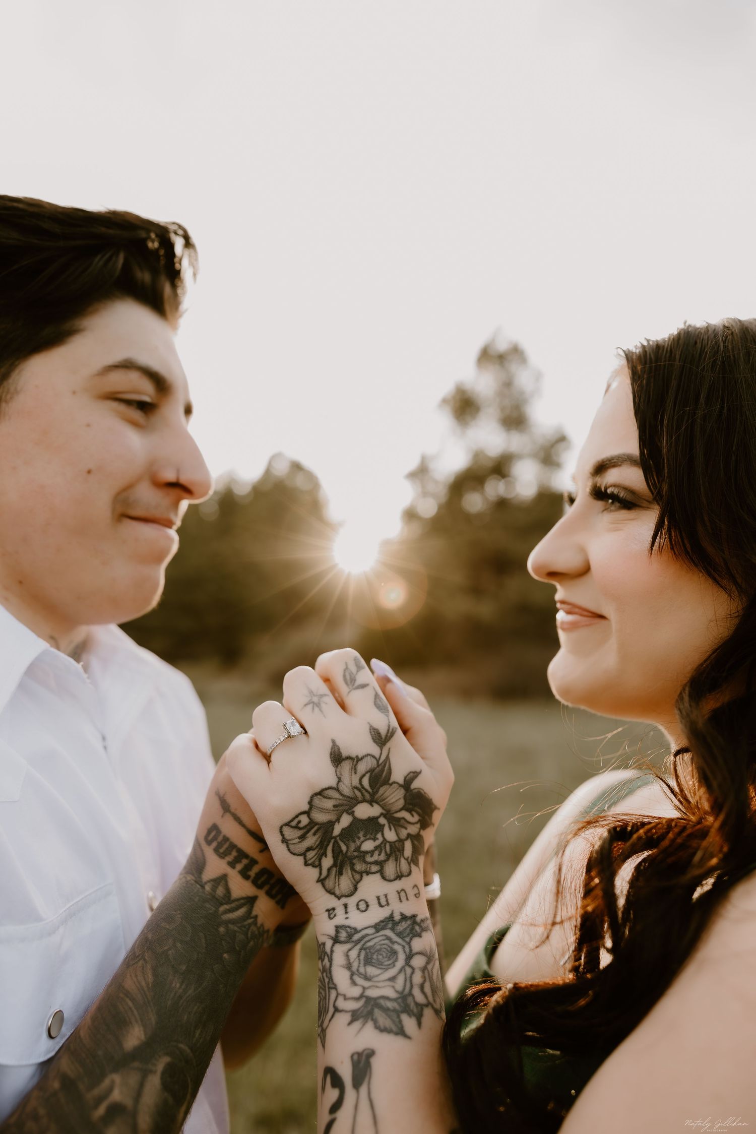 Romantic couple holds hands with visible tattoos during golden hour sunset in outdoor setting.