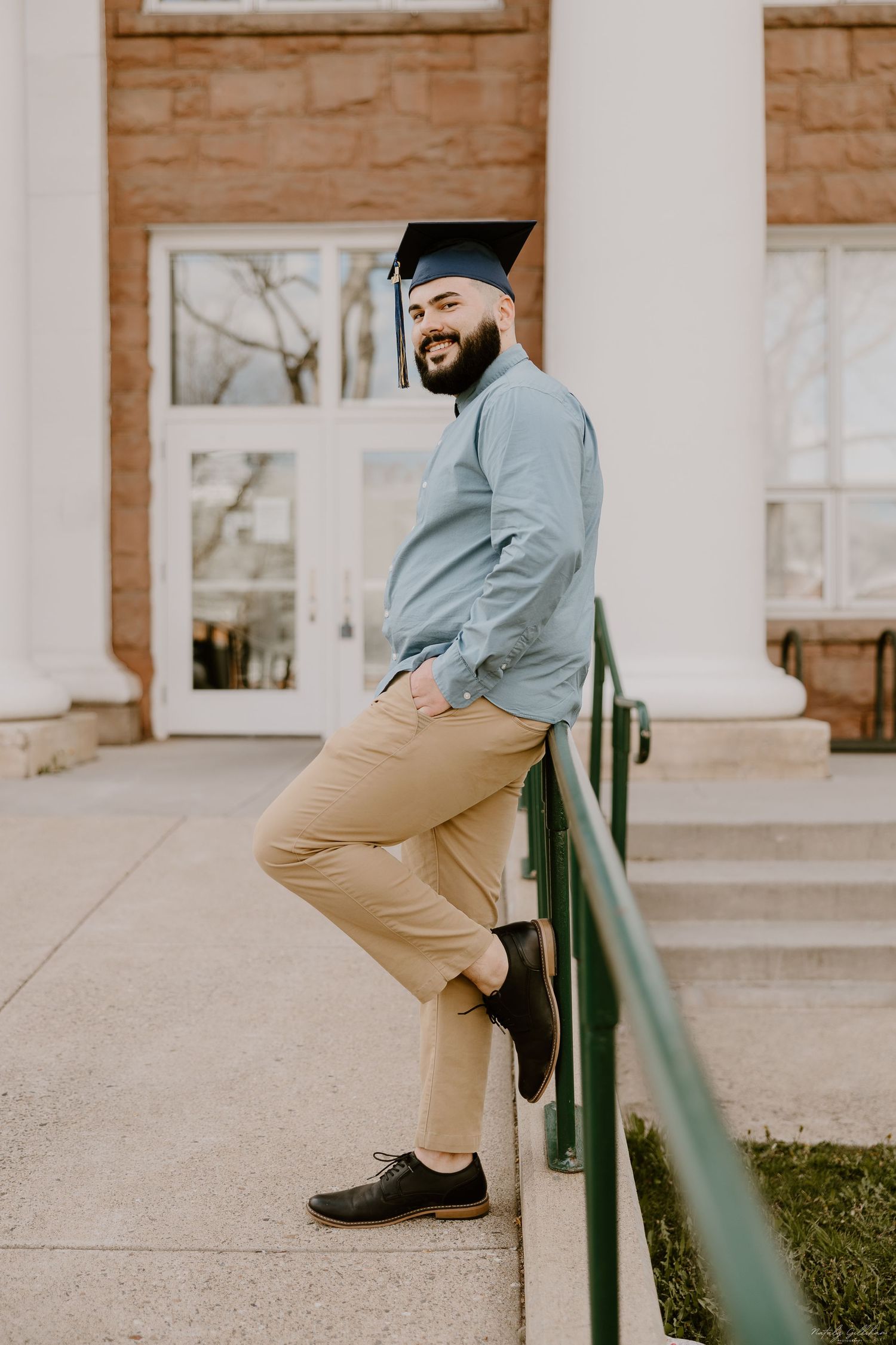 Graduate in academic cap wearing light blue shirt and khaki pants poses casually on building steps with green railing.