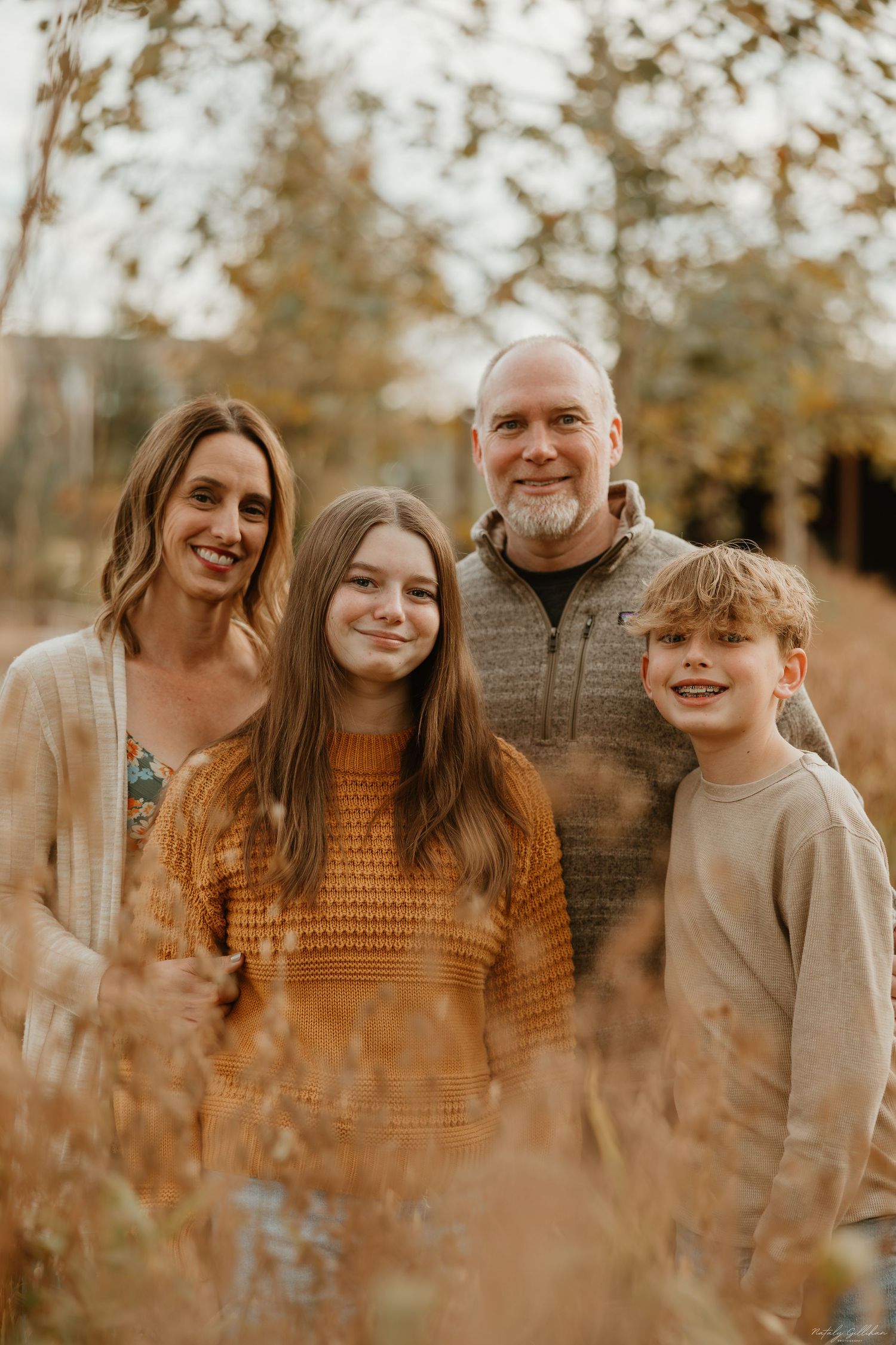 A family of four poses together outdoors on an autumn day with golden grass and fall foliage in the background.