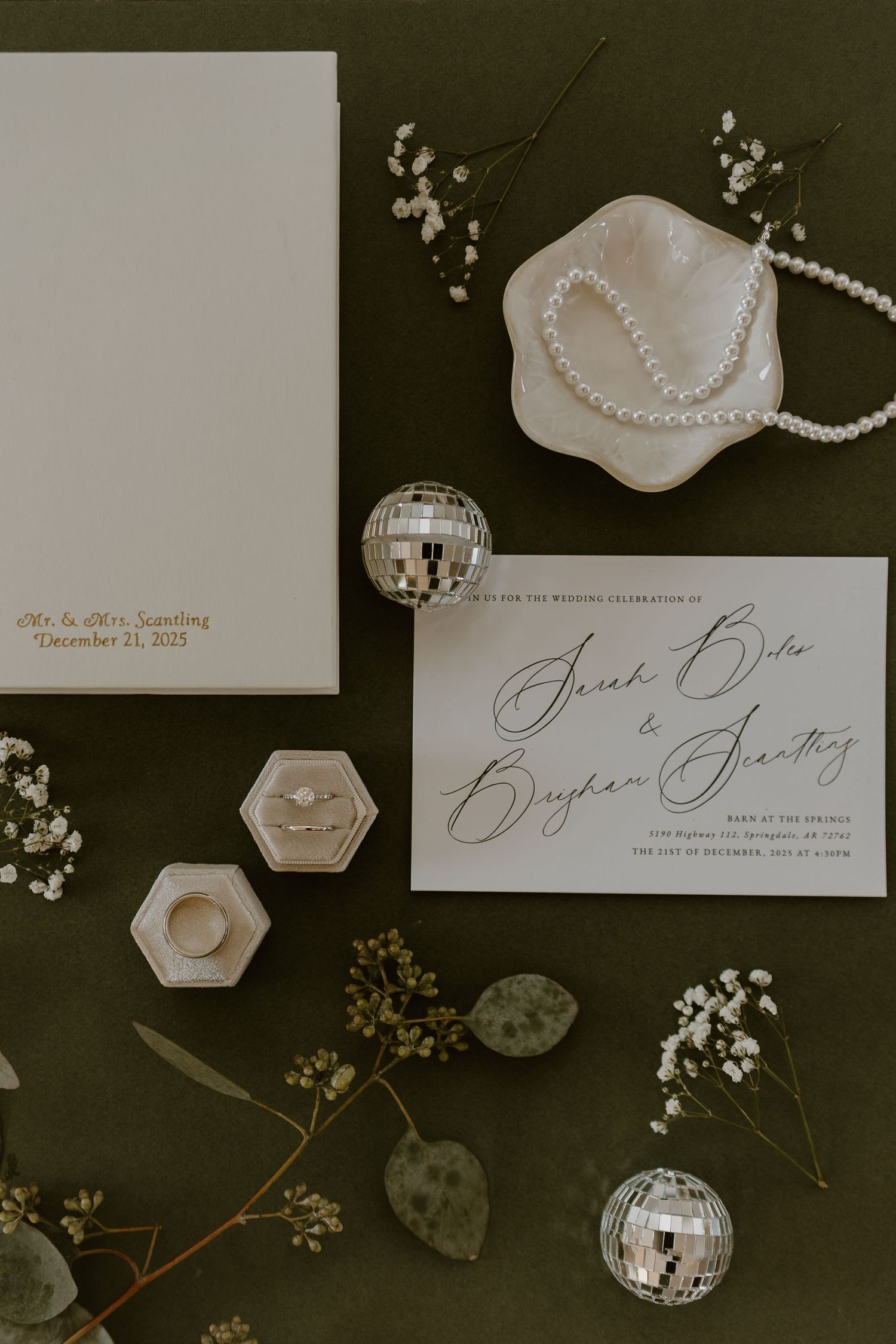 Elegant wedding stationery flatlay with pearl necklace, hexagonal dishes, and delicate floral accents on dark background.