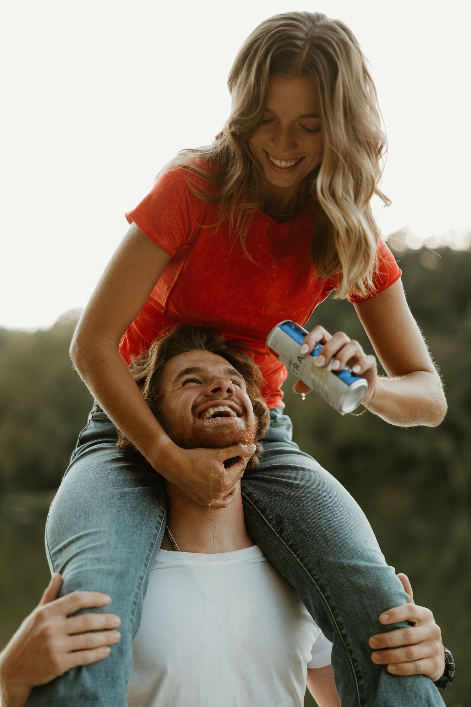 Playful couple in red shirt and white tee laughing together during golden hour outdoor photoshoot.