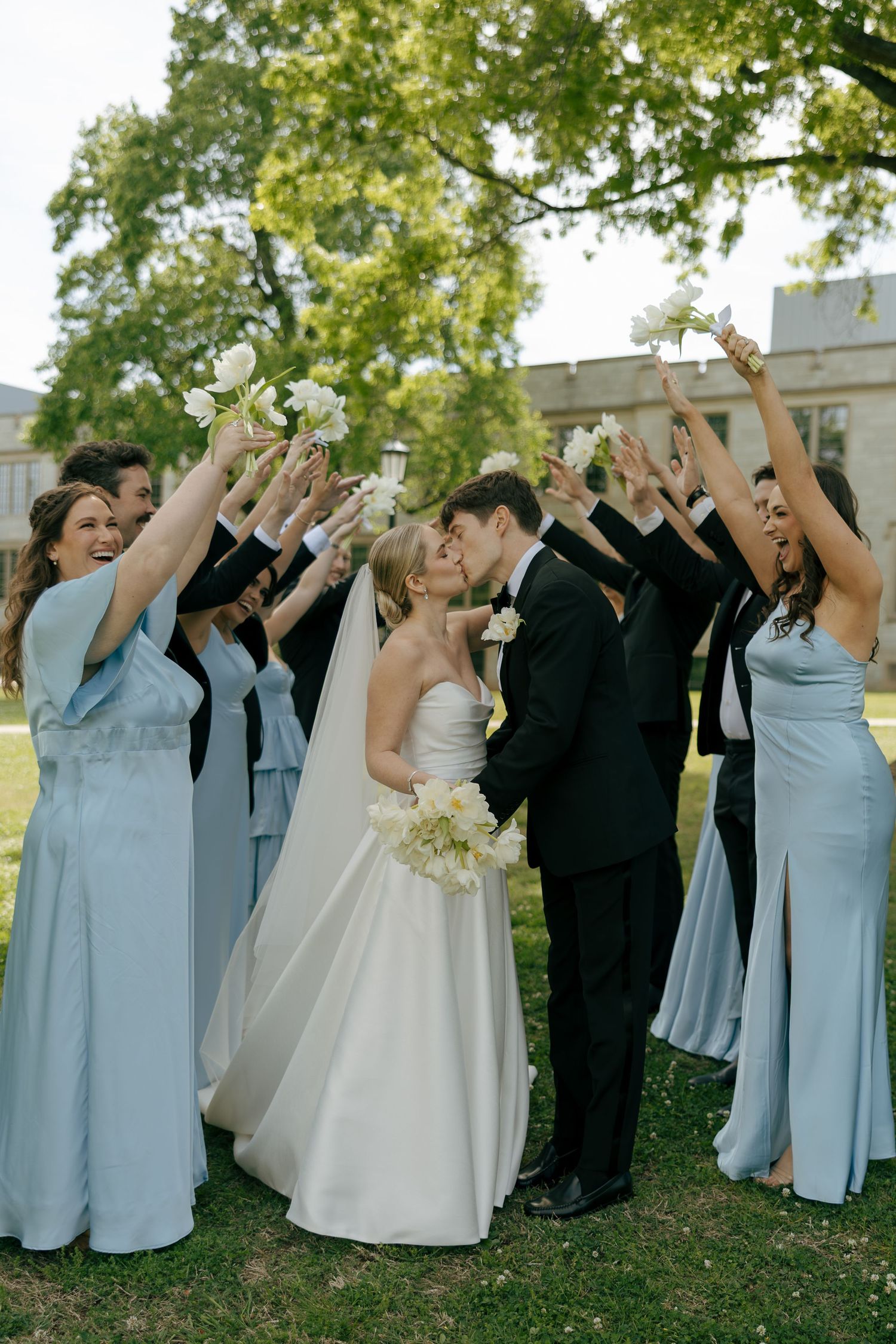 Bride and groom share a kiss surrounded by bridesmaids in blue gowns and groomsmen raising bouquets outdoors.
