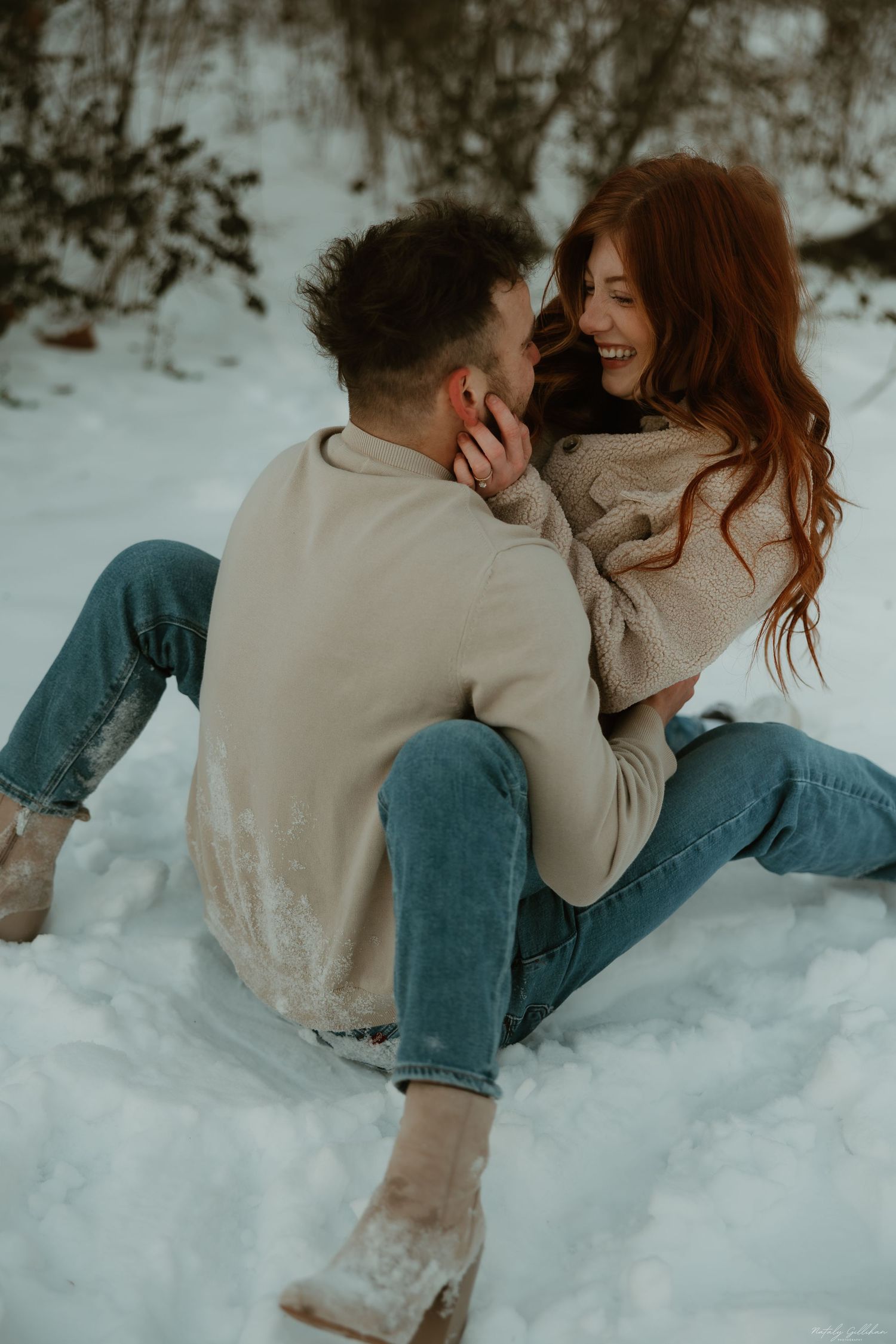 Couple in matching beige sweaters laughing together while sitting in snowy winter setting.