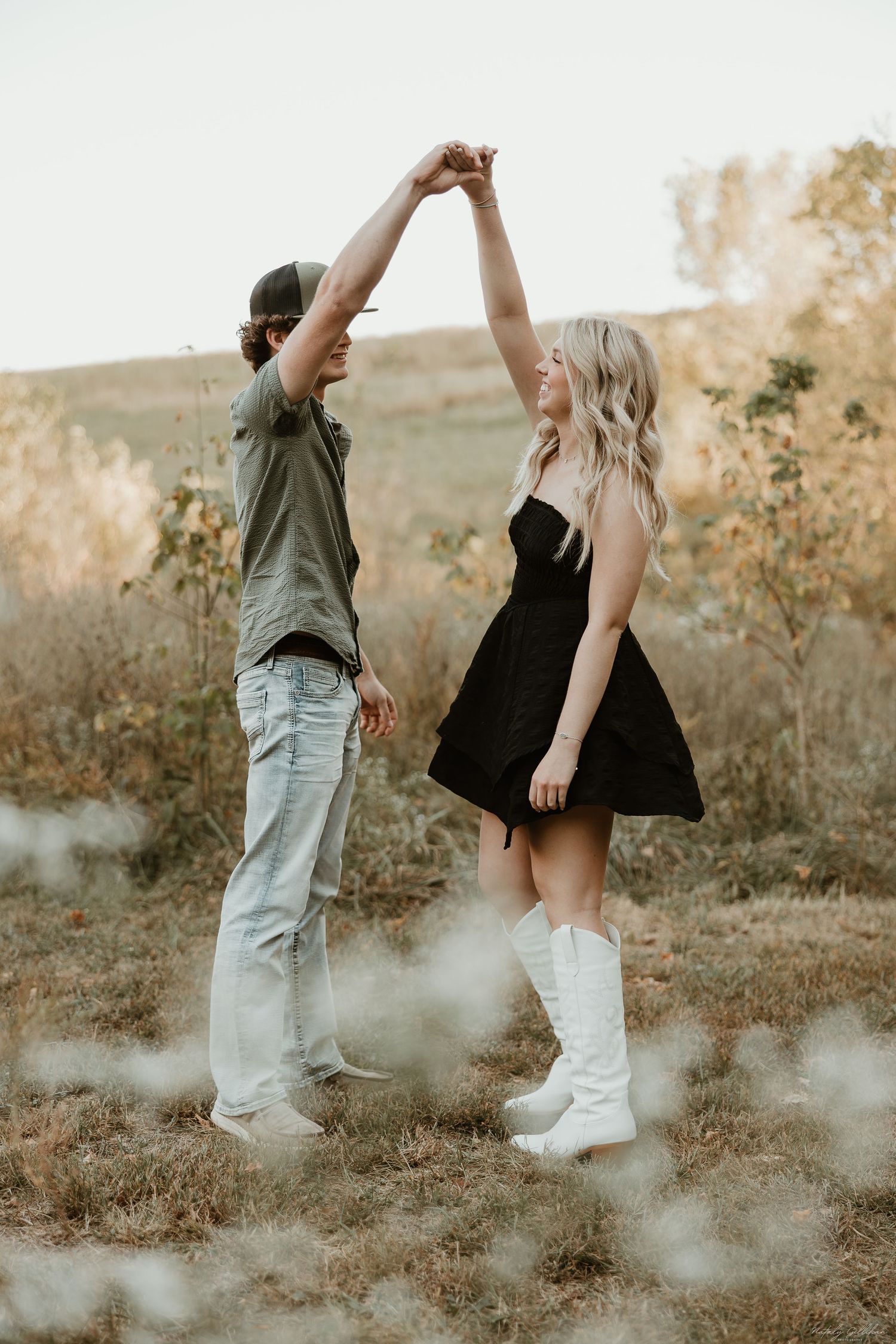 A couple dances together on a hillside at sunset, with the woman wearing a black dress and white boots.