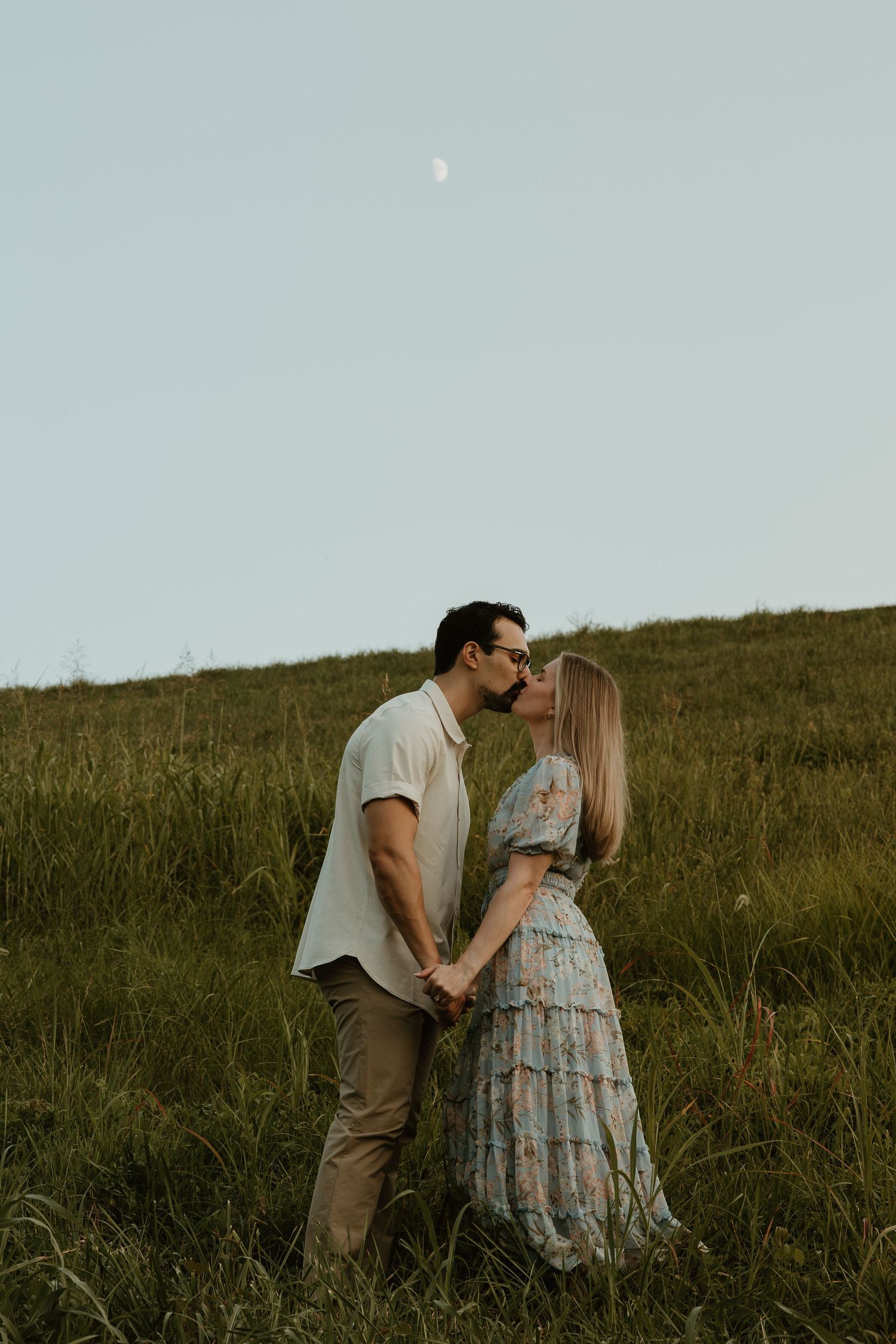 A romantic couple embraces on a grassy hillside under a pale dusk sky with a visible moon.