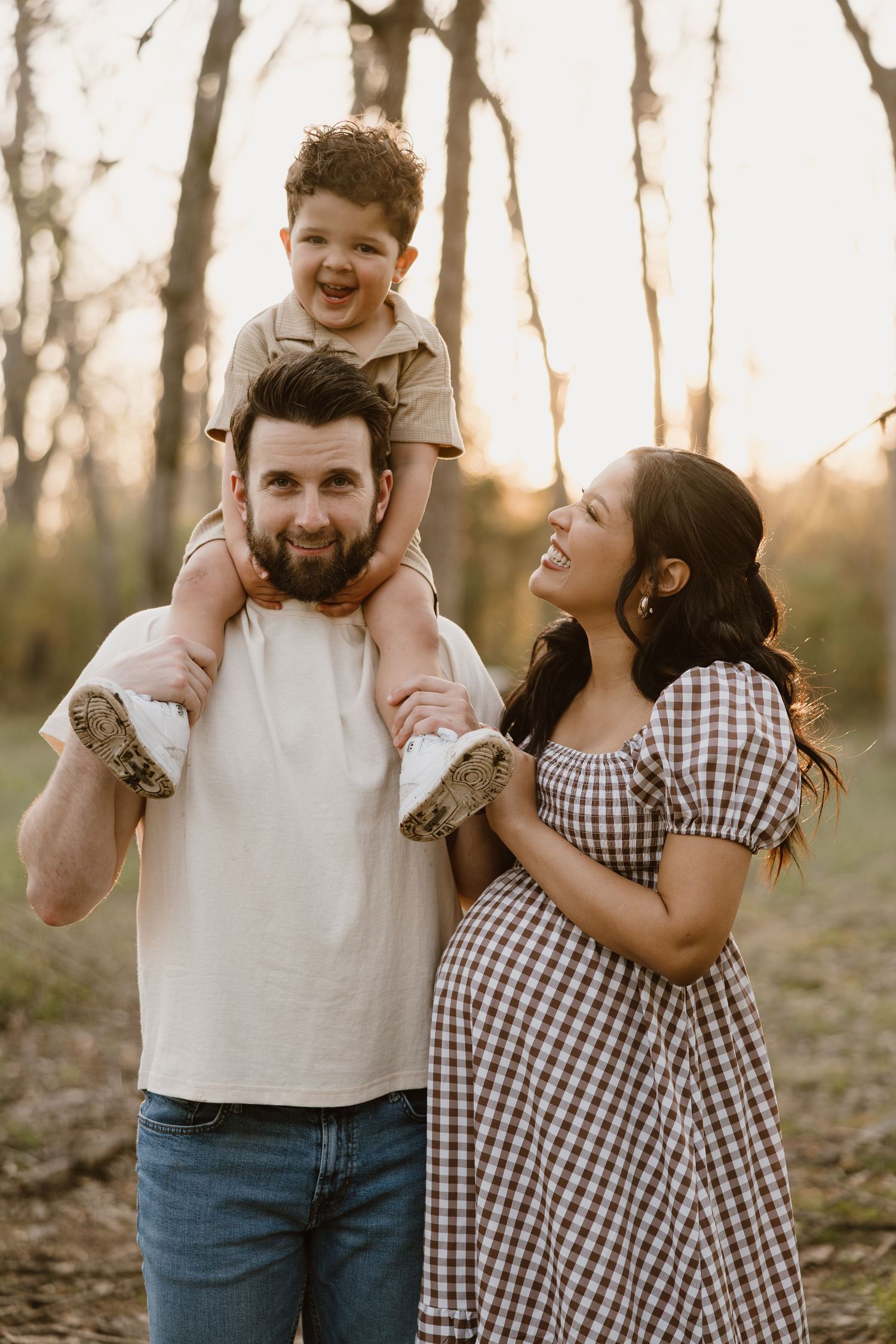 Happy family outdoors at sunset, father carrying toddler on shoulders, pregnant mother in gingham dress smiling.