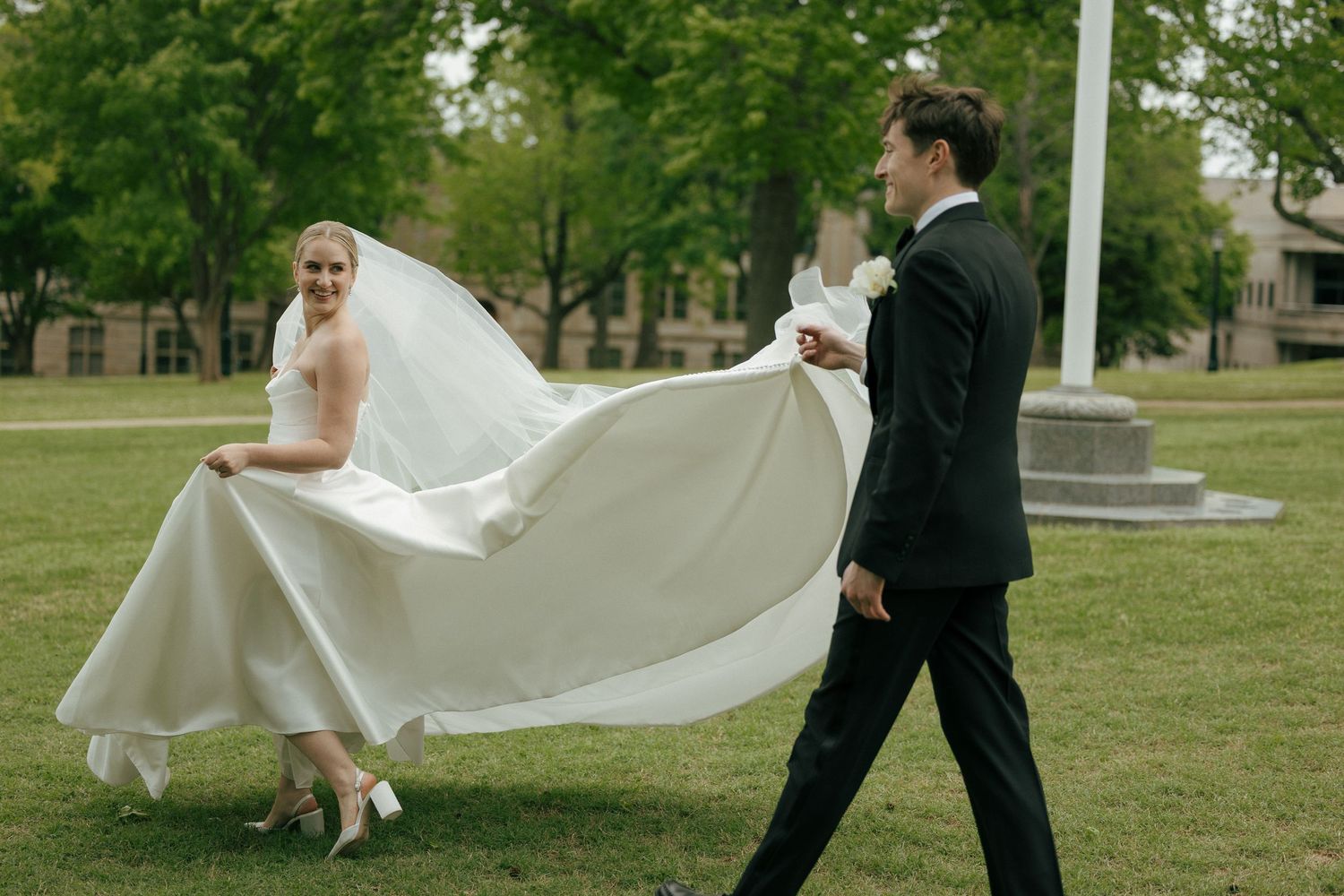 Bride in flowing white gown and veil walks with groom in black suit across green lawn at outdoor wedding.