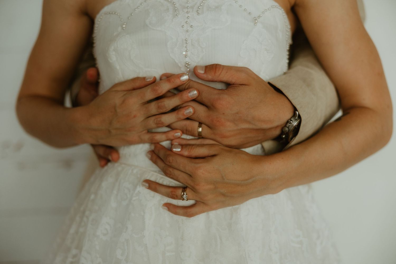 Close-up of hands with wedding rings resting on white lace wedding dress showing romantic wedding details.