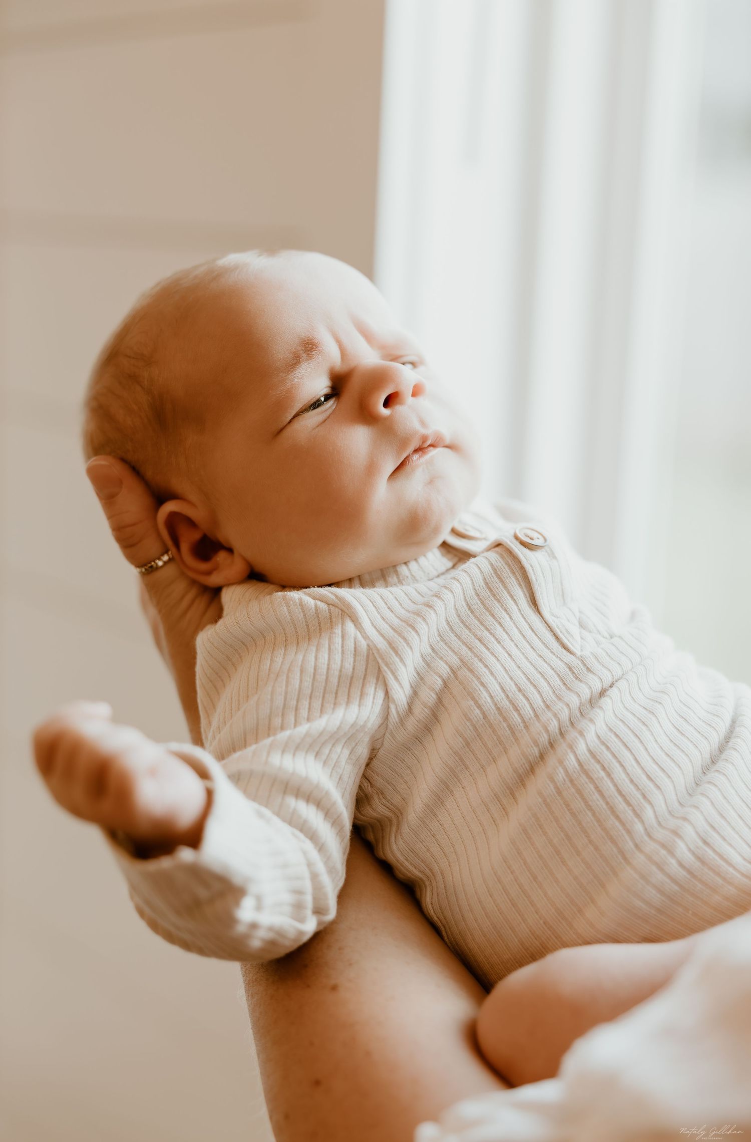 A peaceful newborn baby wearing a white ribbed onesie rests in gentle natural window light.