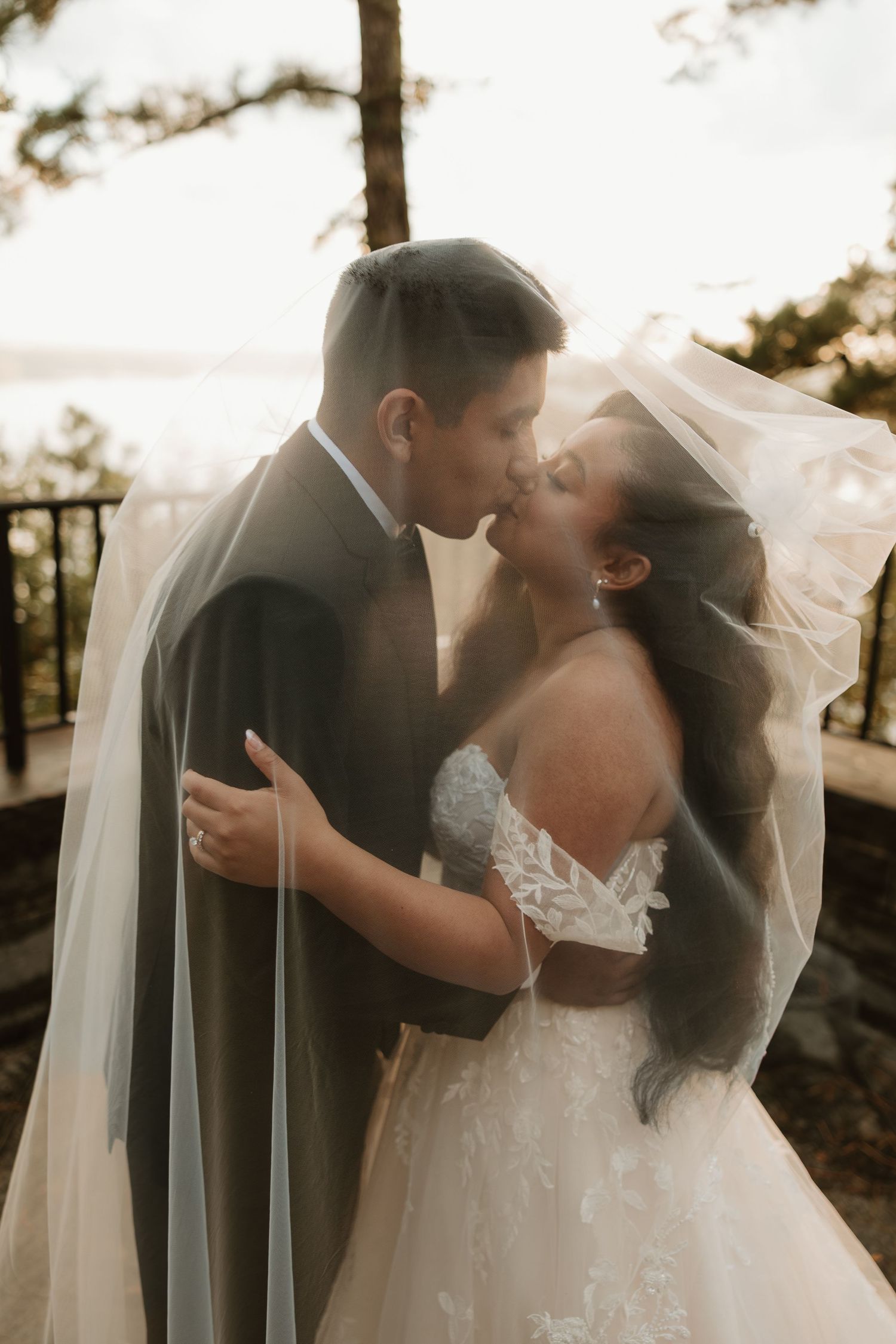 Romantic wedding couple embraces under flowing veil during sunset outdoor photo session.