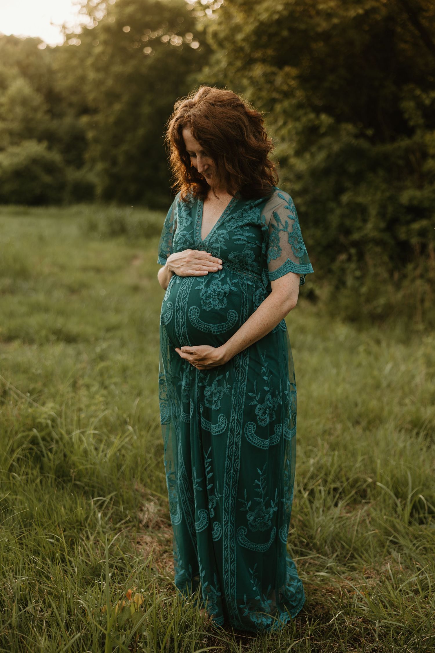 Expectant mother in flowing teal lace dress cradles her baby bump while standing in a sunlit meadow at golden hour.