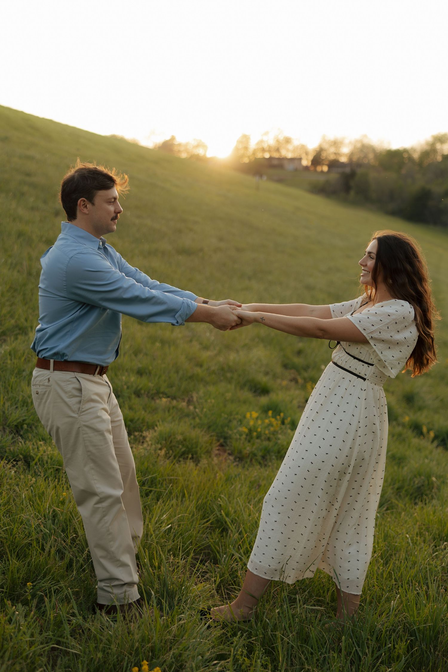 Couple holding hands and dancing in a green field at golden hour sunset, woman in white floral dress.