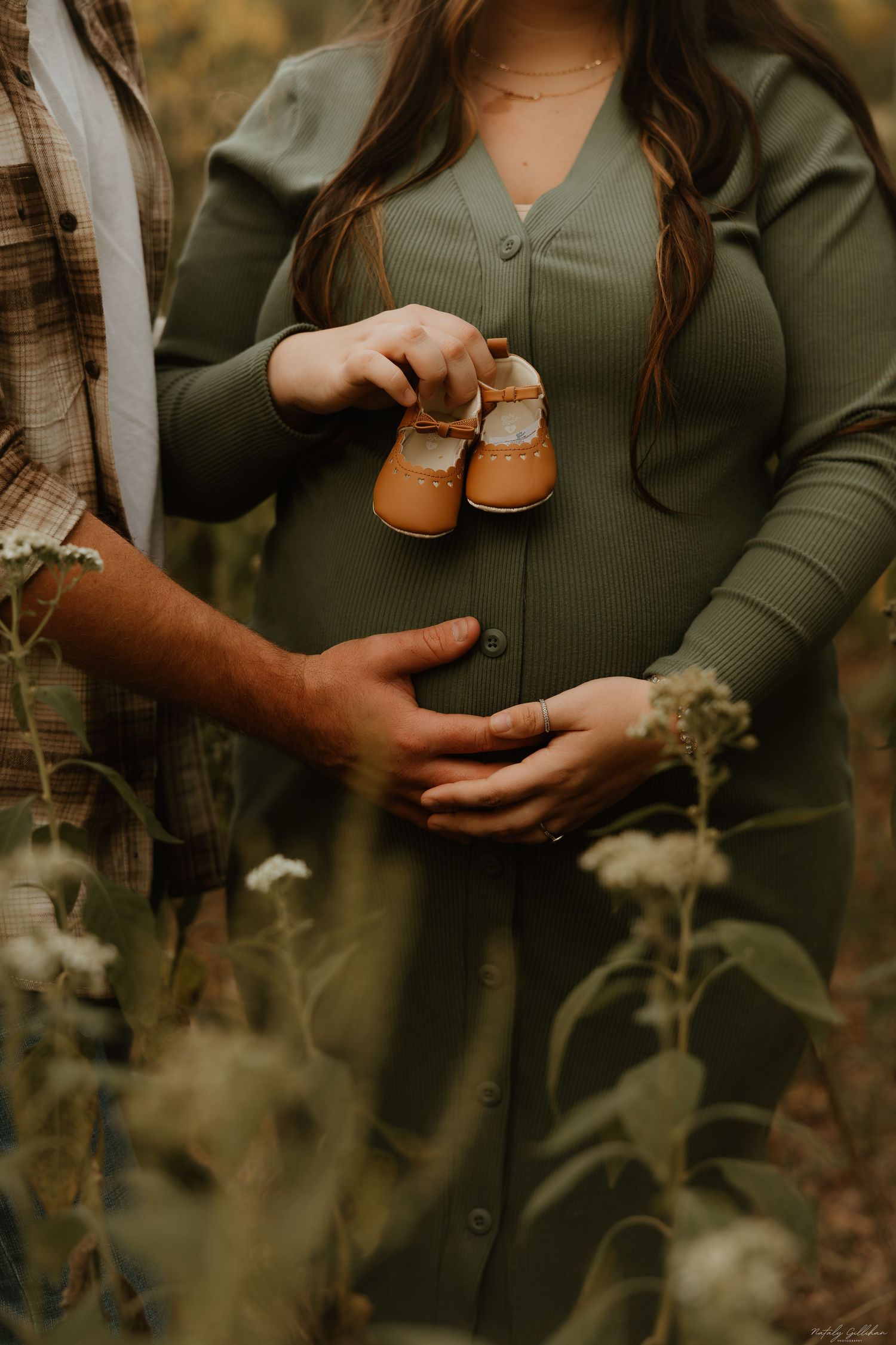 Hands hold tiny brown baby shoes against a green knit maternity dress in an outdoor autumn setting.