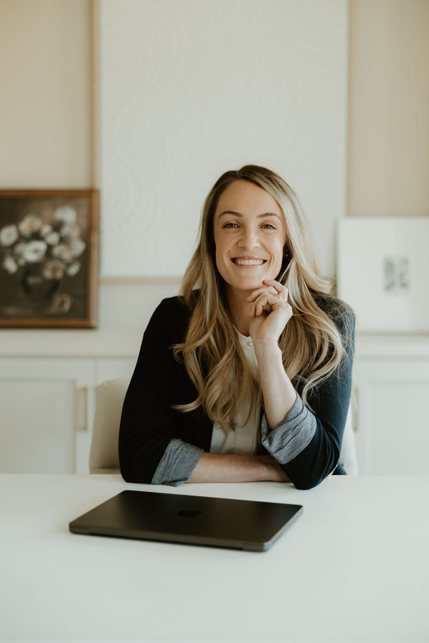 Professional woman with blonde hair smiles warmly while sitting at desk with tablet in elegant office interior.