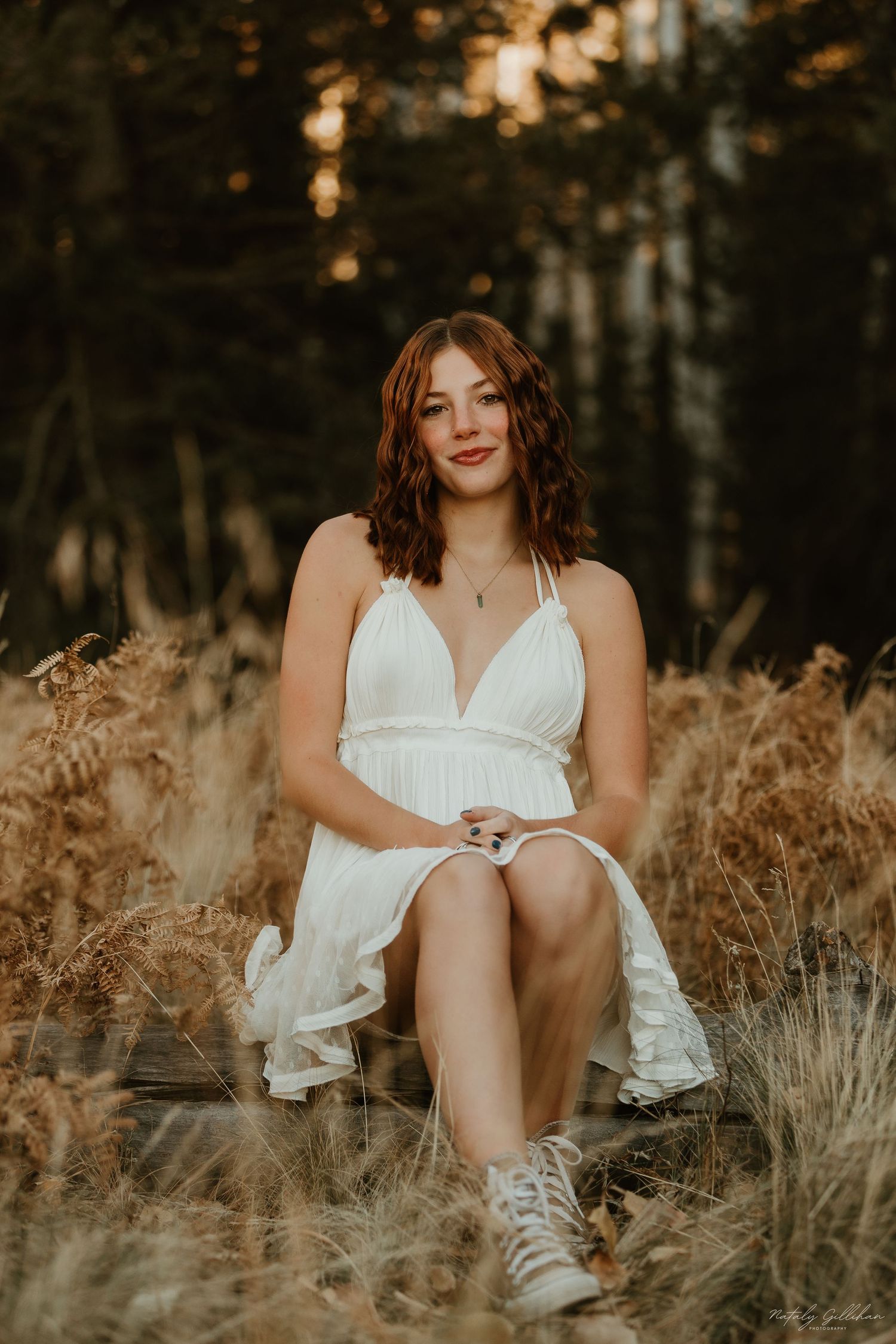 Someone in a flowing white dress and sneakers sits among golden dried grasses at sunset in a moody forest setting.