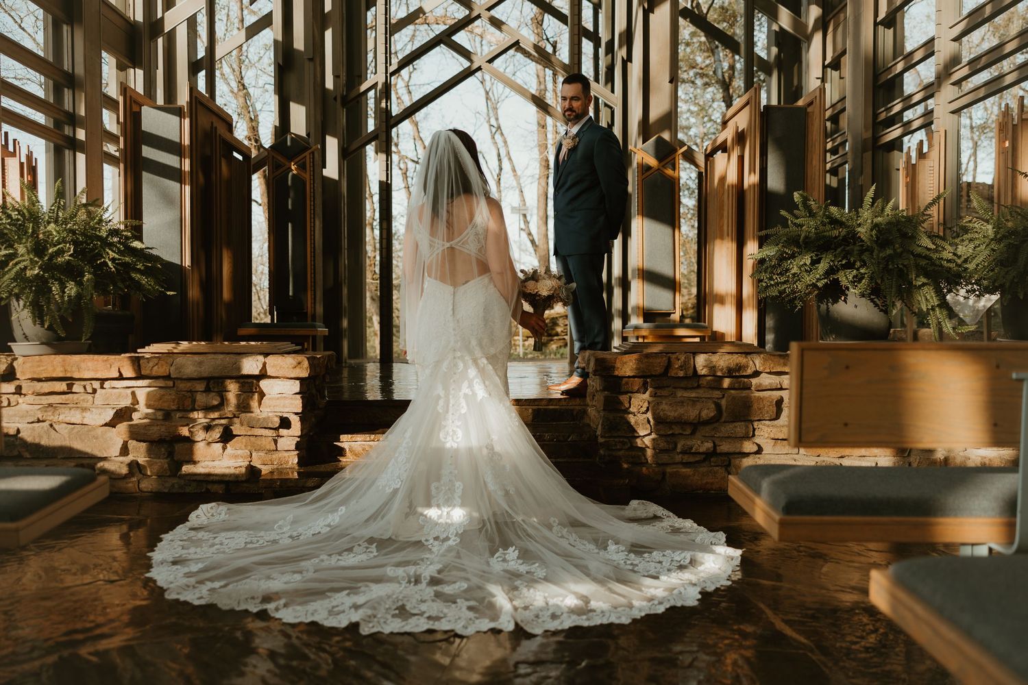 Bride and groom stand in elegant glass chapel with dramatic long veil trailing across stone floor during wedding ceremony.