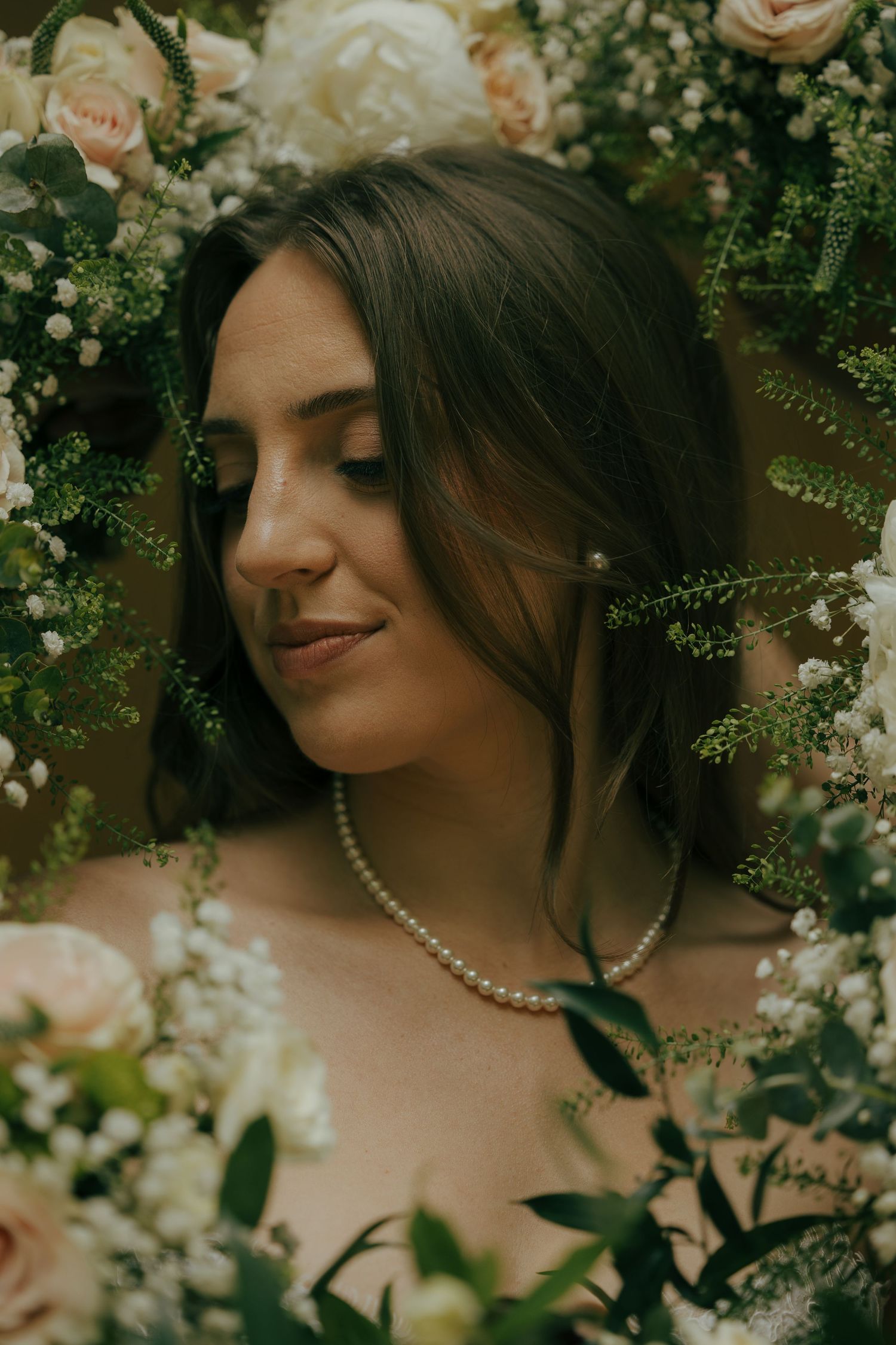 Serene bride in pearl necklace surrounded by lush white and blush floral arrangements.