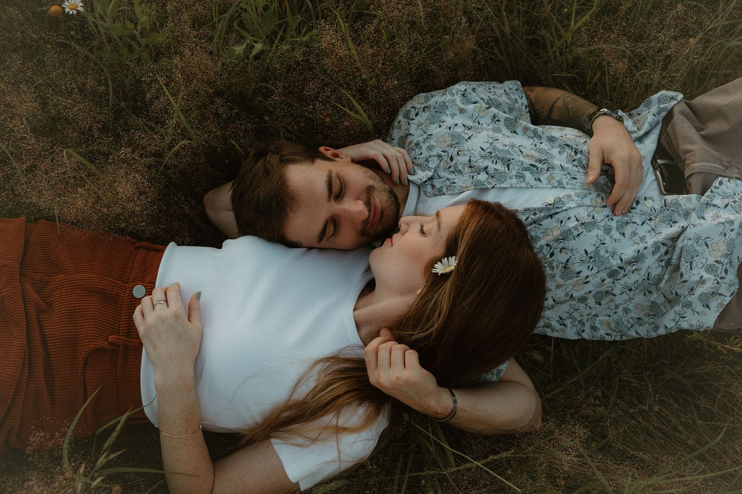 Two people in casual attire lying in grass together on warm brown earth during a golden sunset.