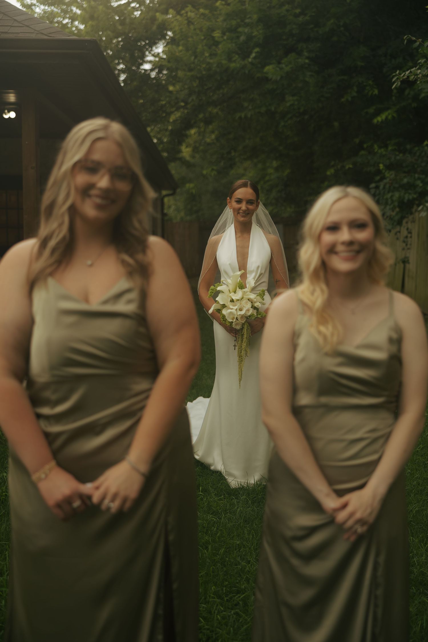 Bride in white gown with veil holds bouquet while two bridesmaids in olive satin dresses stand in foreground outdoors.