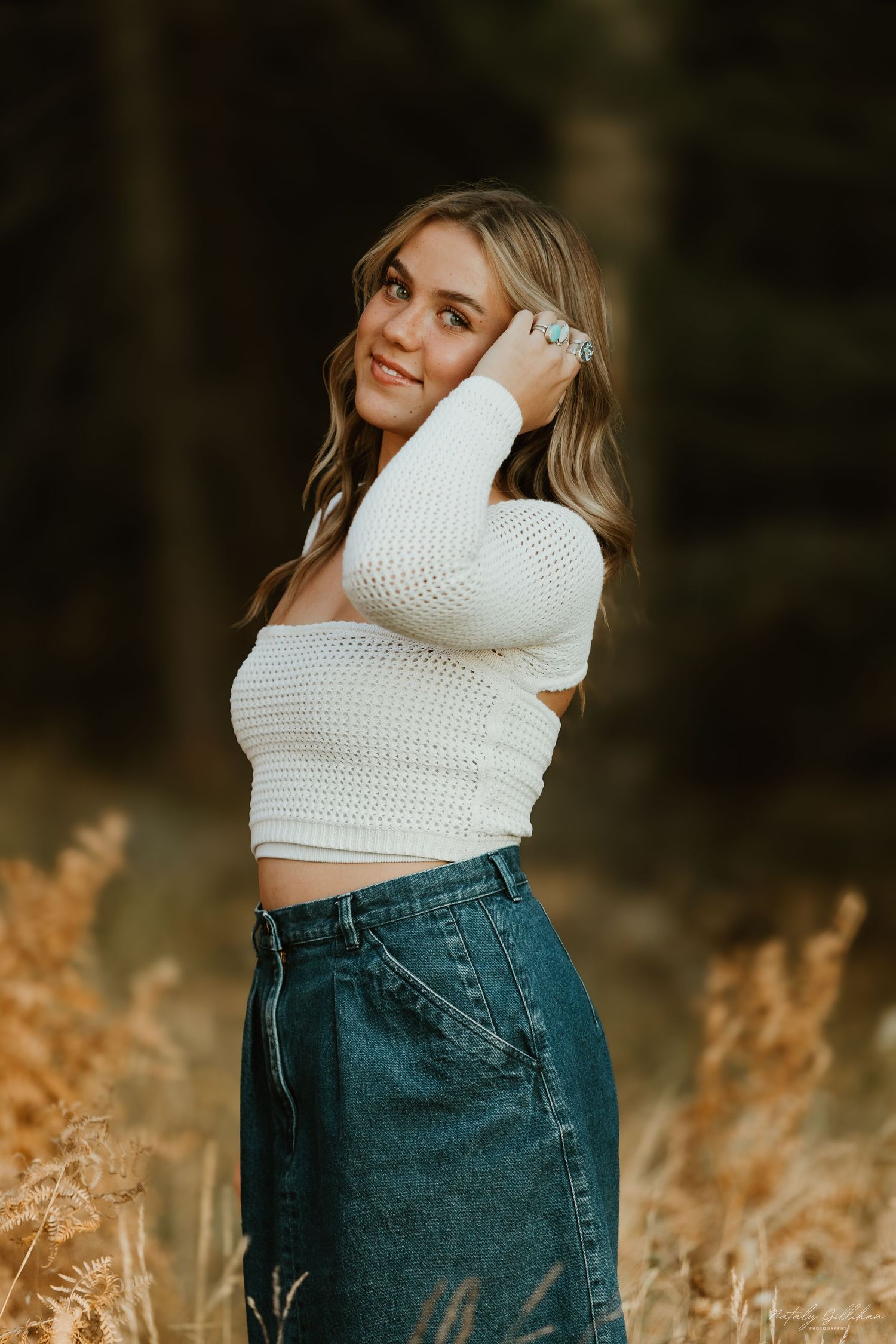 Woman wearing white knit crop top and denim skirt poses in golden field during sunset.