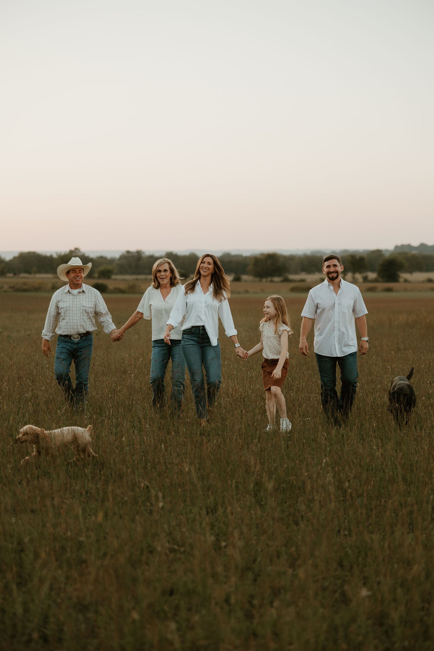 Family of five walking through a grassy field at dusk with two dogs, wearing white shirts and jeans.