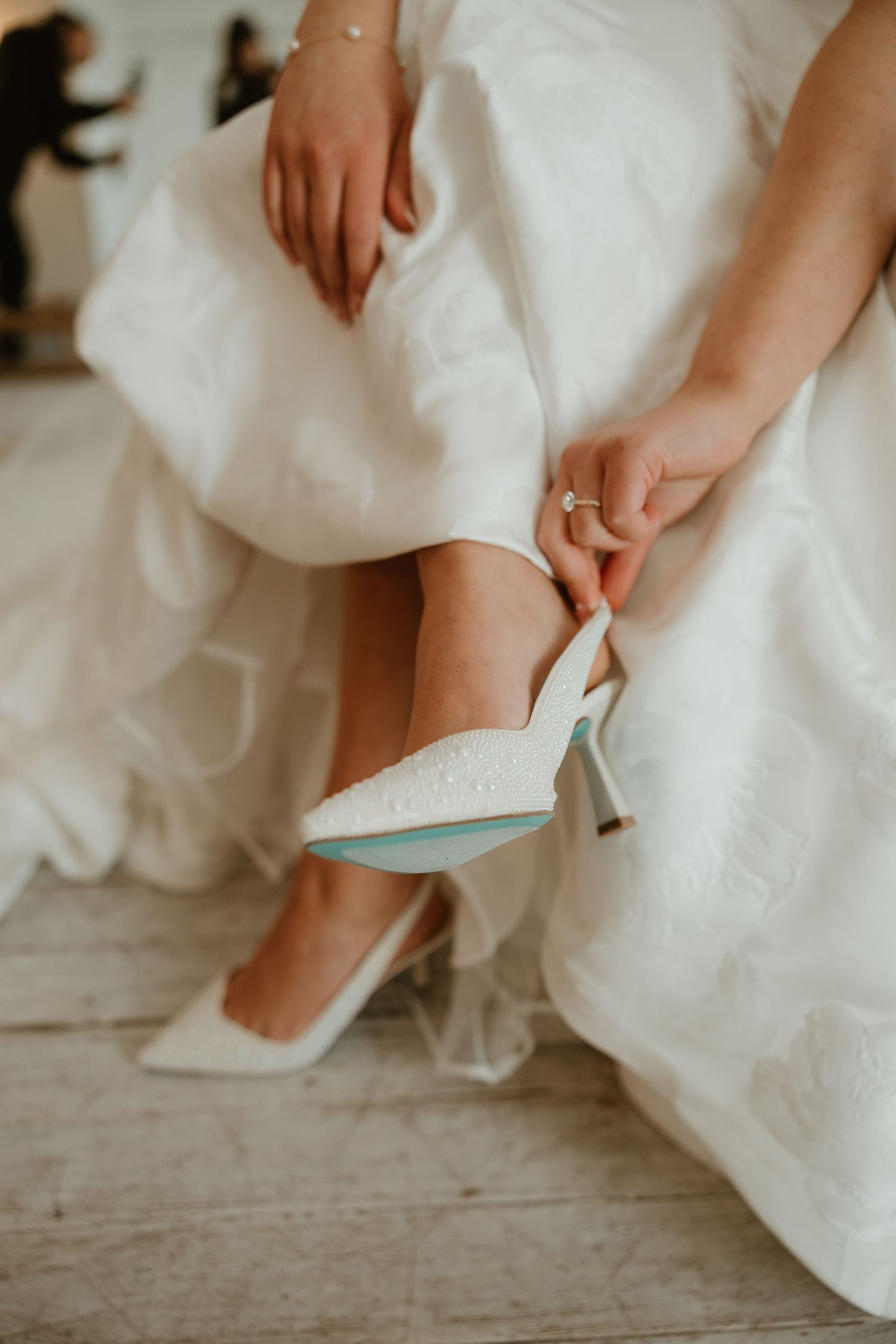 A close-up shot of hands adjusting an elegant white wedding shoe against a flowing white dress and wooden floor.