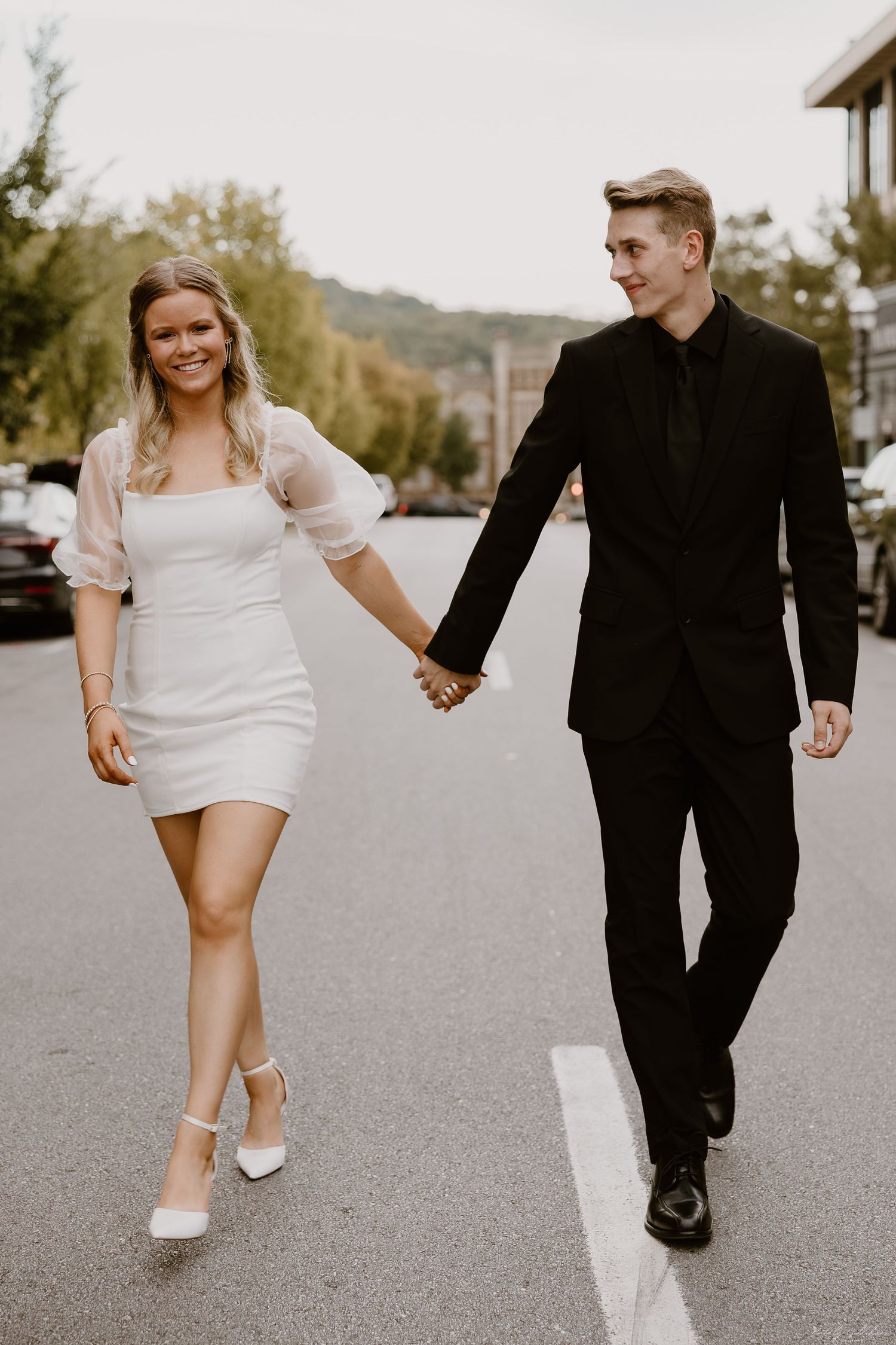 Couple walks down street holding hands, wearing black suit and white dress.