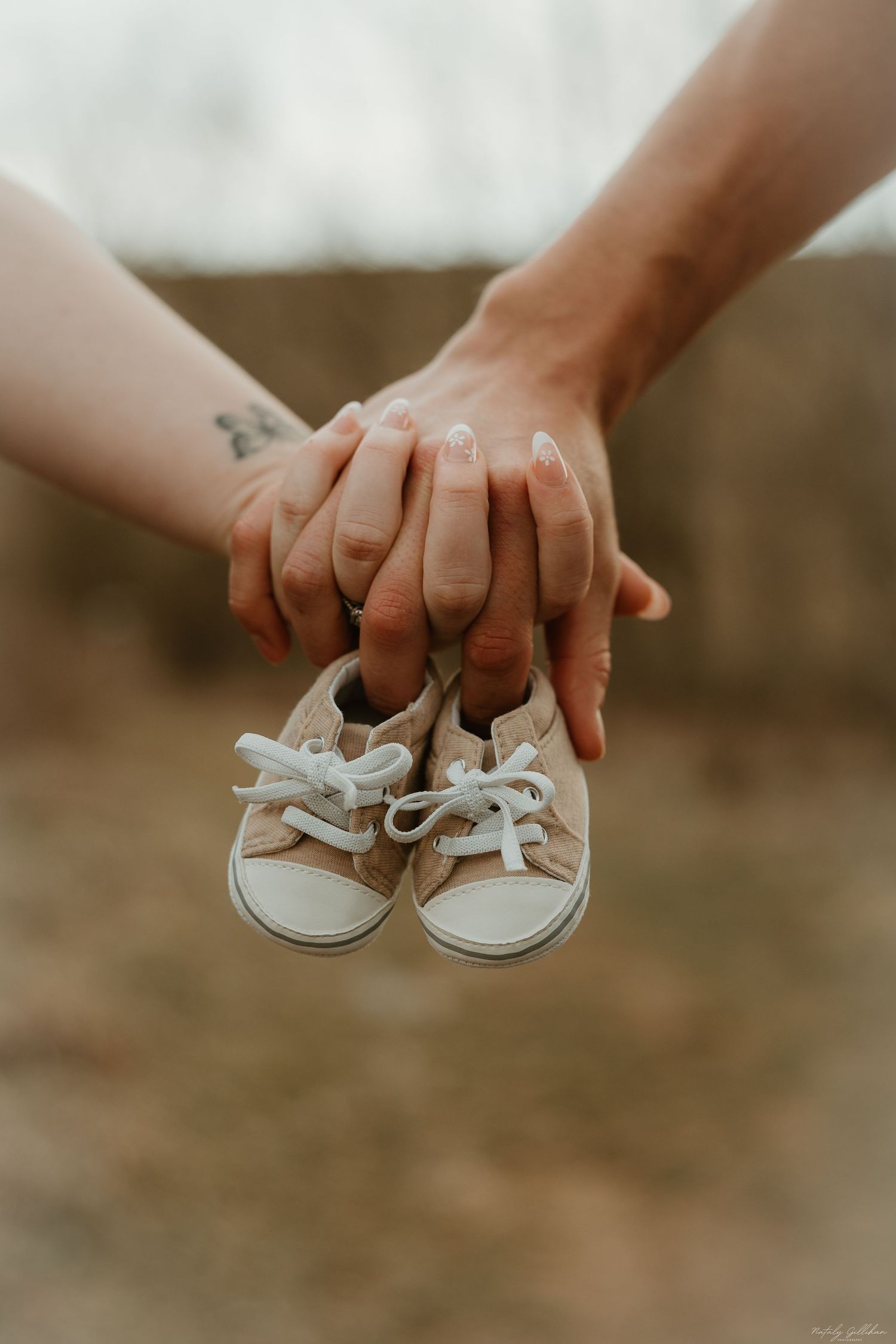 Two hands holding small beige baby shoes against a blurred outdoor background in warm muted tones.