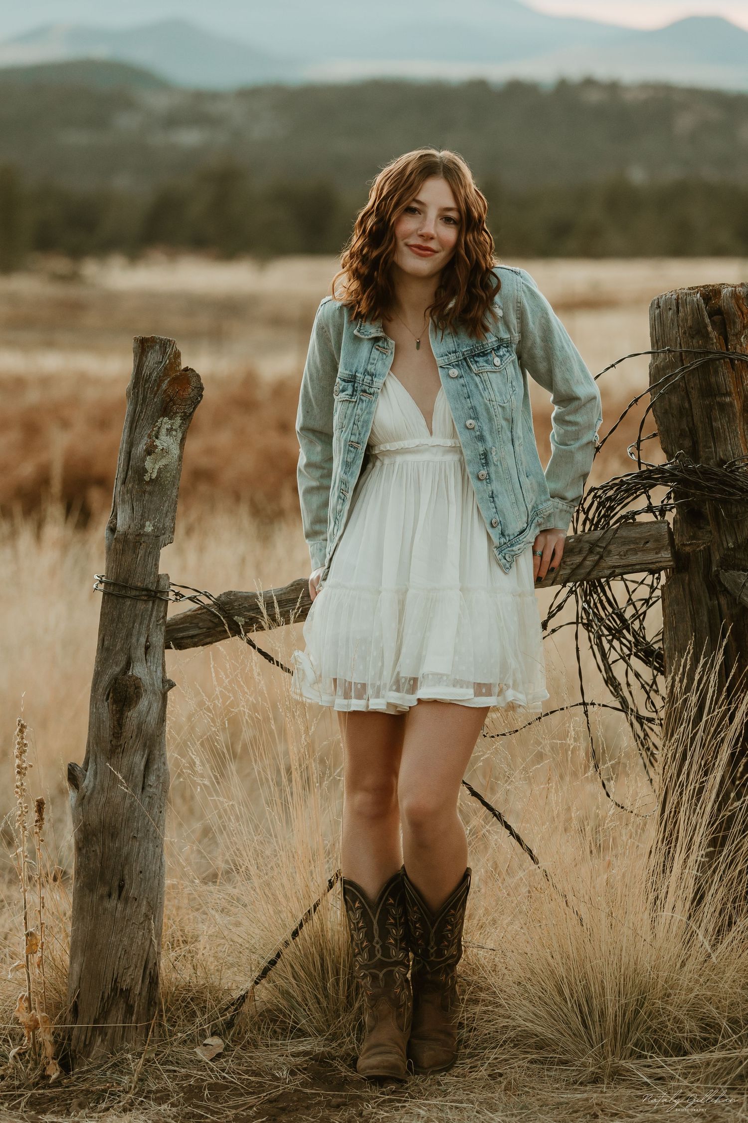 A rustic country portrait in a field showing cowboy boots, white dress and denim jacket against mountain backdrop.