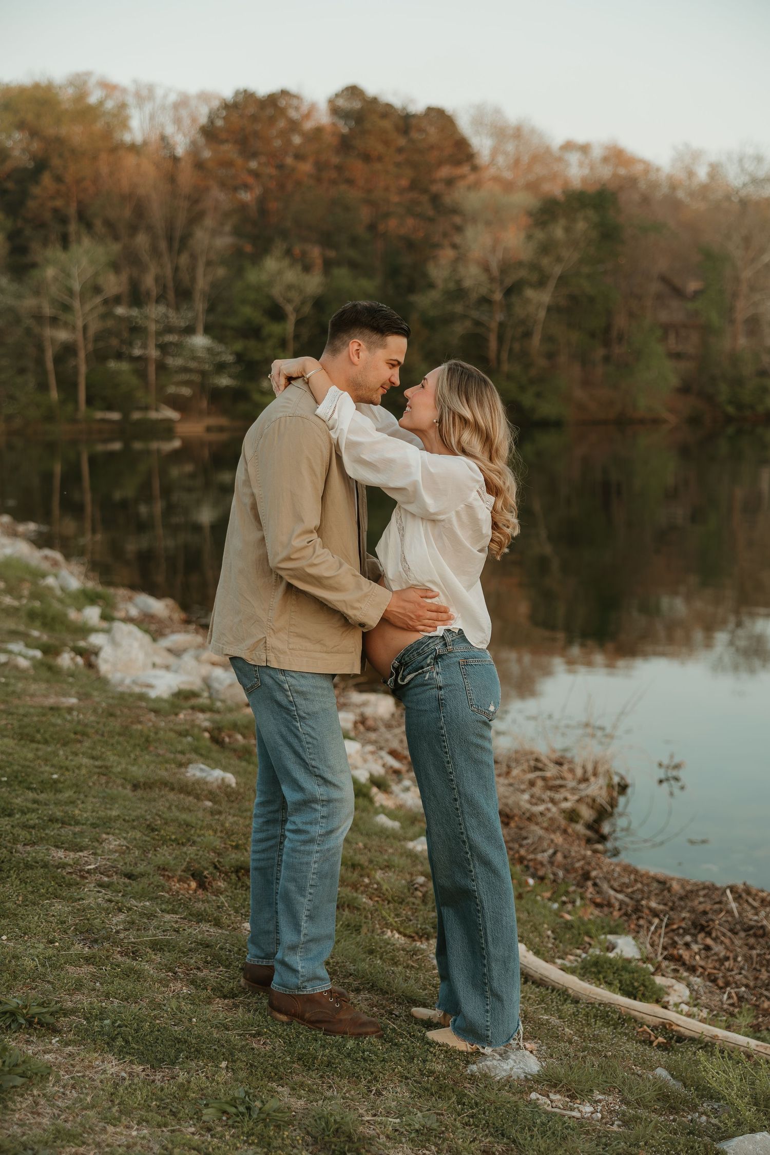 Couple embracing and almost kissing by a serene lake with autumn trees reflected in the water.