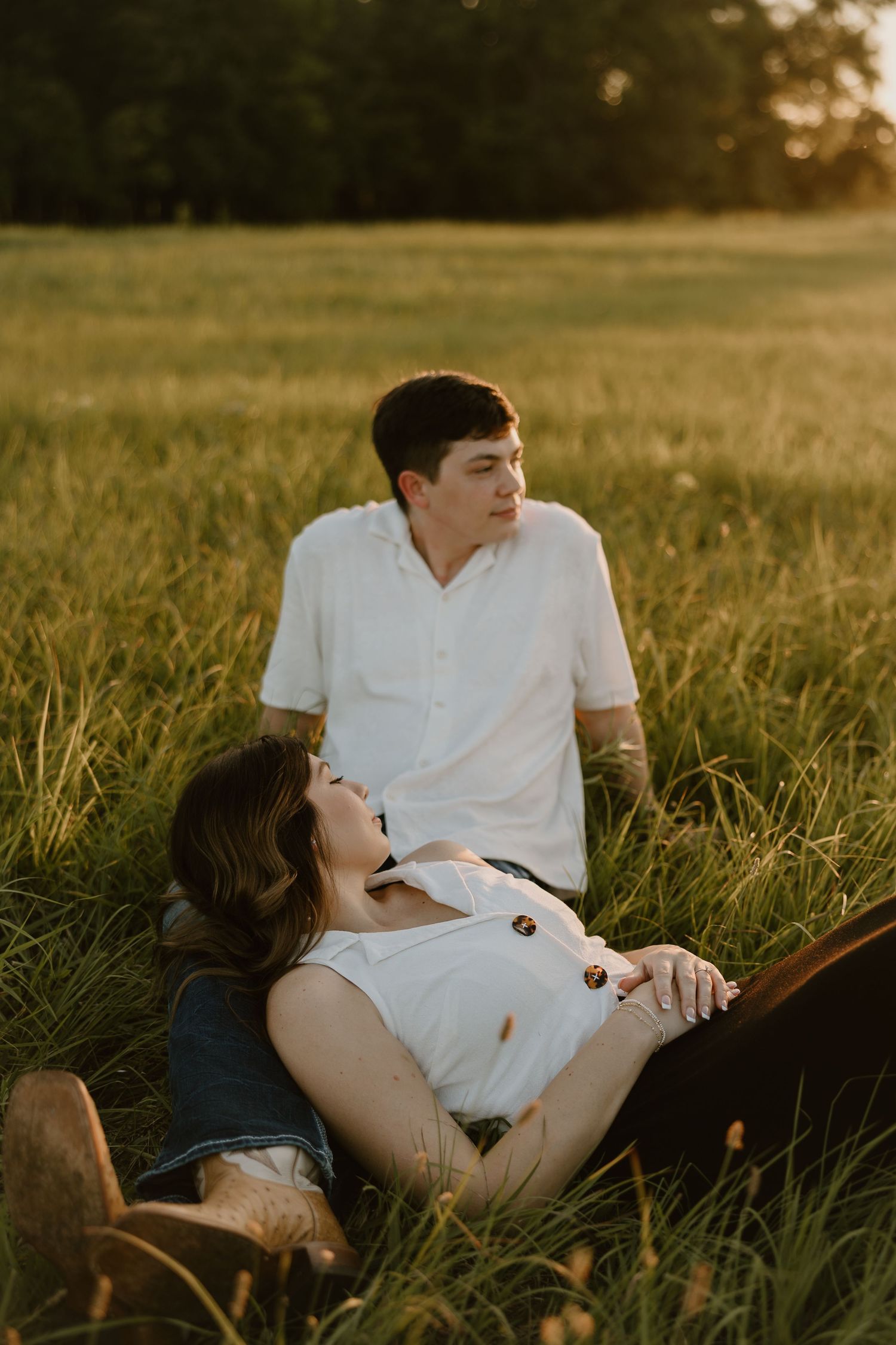 A couple in white shirts lies in a golden sunlit meadow during sunset creating an intimate romantic moment.