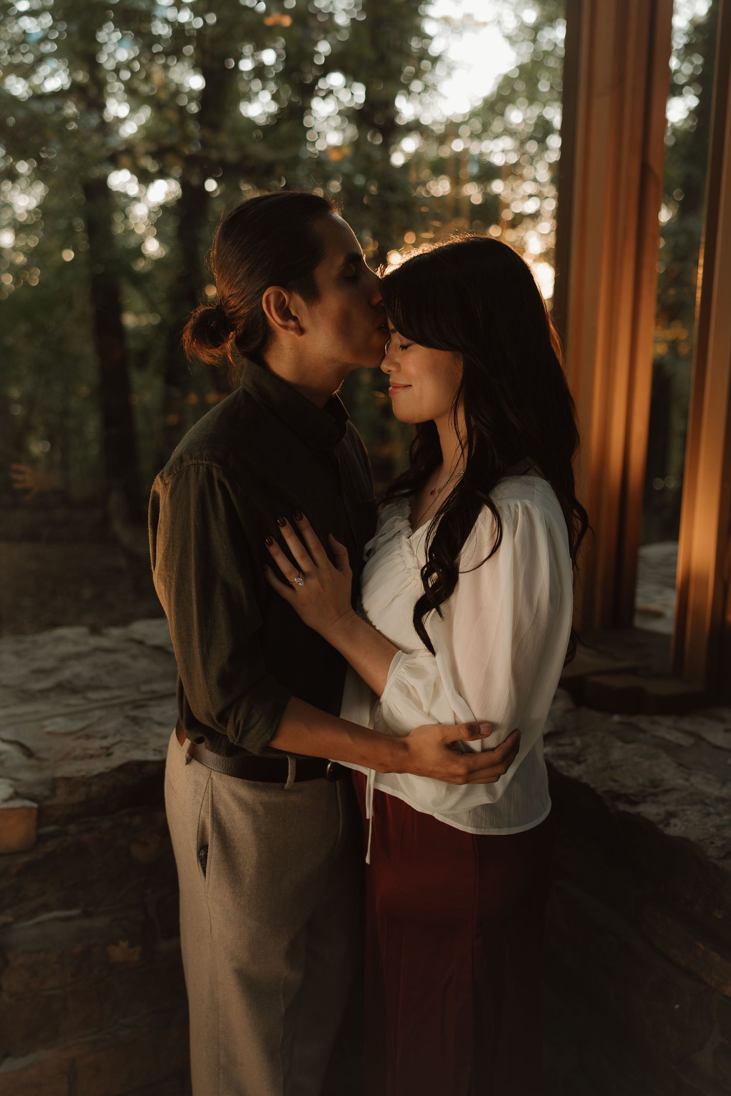 Couple sharing a tender kiss at golden hour near forest cabin, woman in white blouse and man in olive shirt embracing.