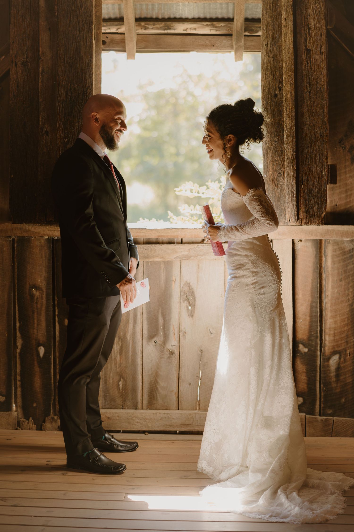 A couple in wedding attire reads letters to each other in a rustic barn setting with natural light streaming through windows.