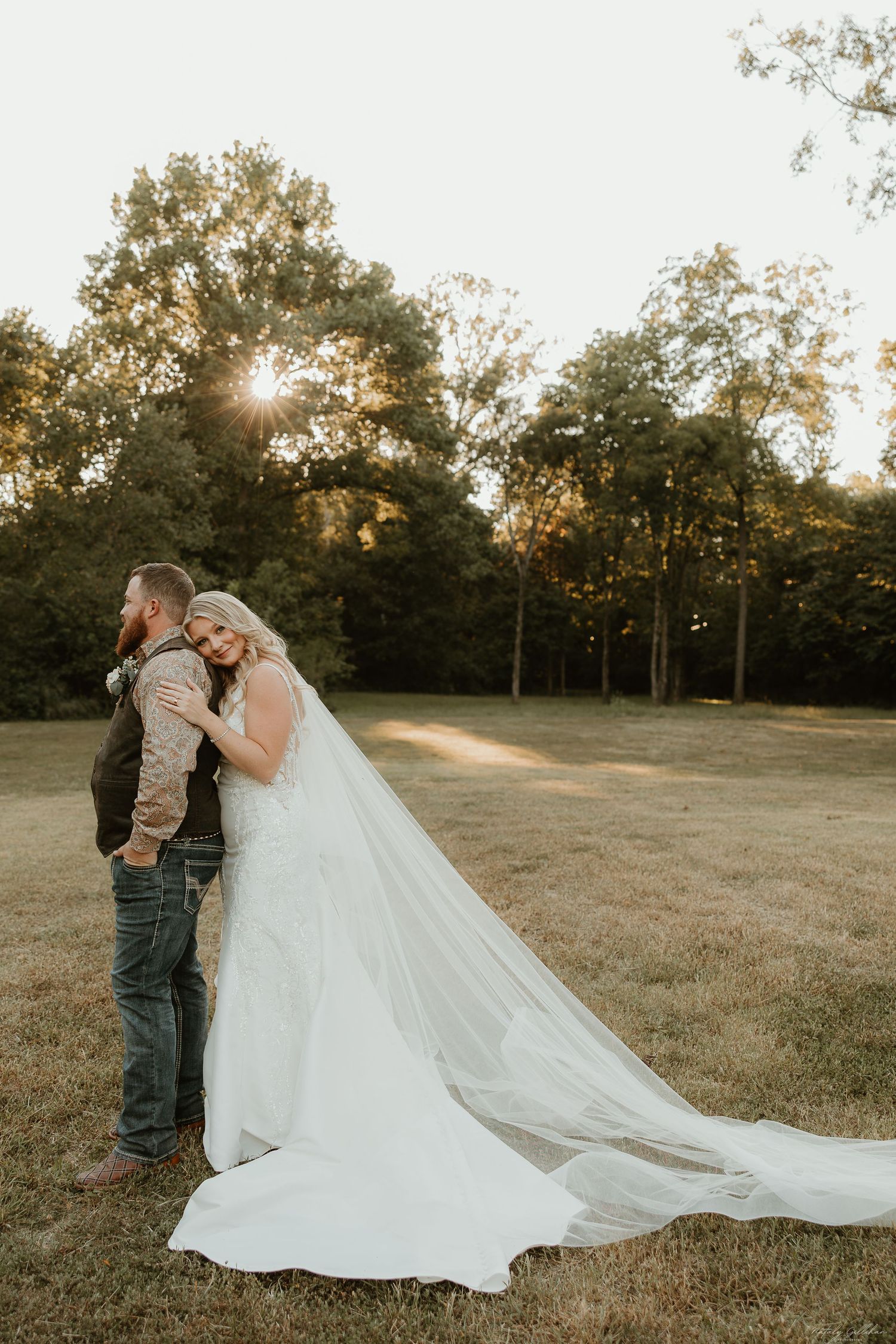 A couple shares an intimate moment at sunset in a field with a flowing white wedding dress and veil.